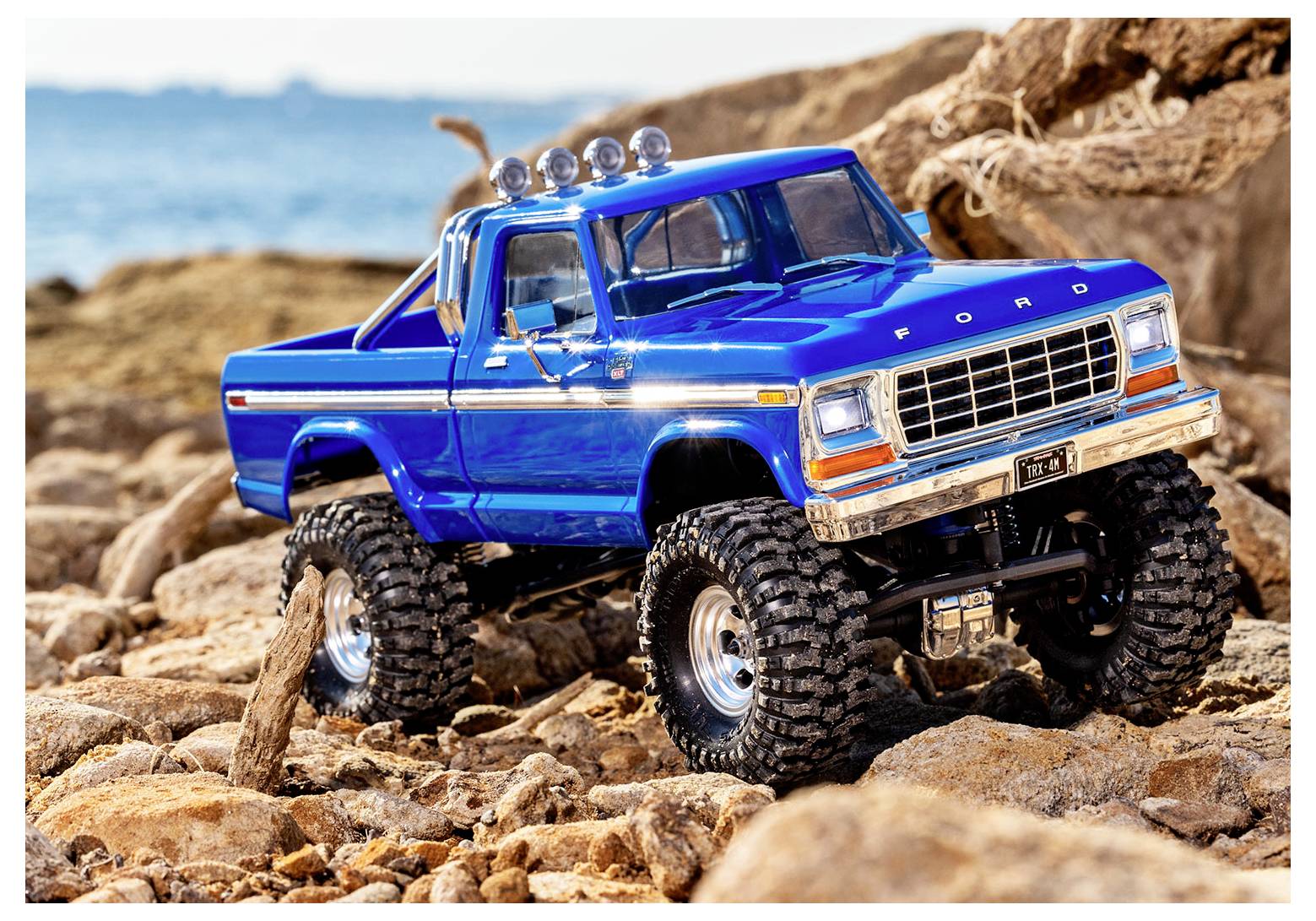 A blue toy truck with large tires sits on rocky terrain, with a blurred ocean in the background.