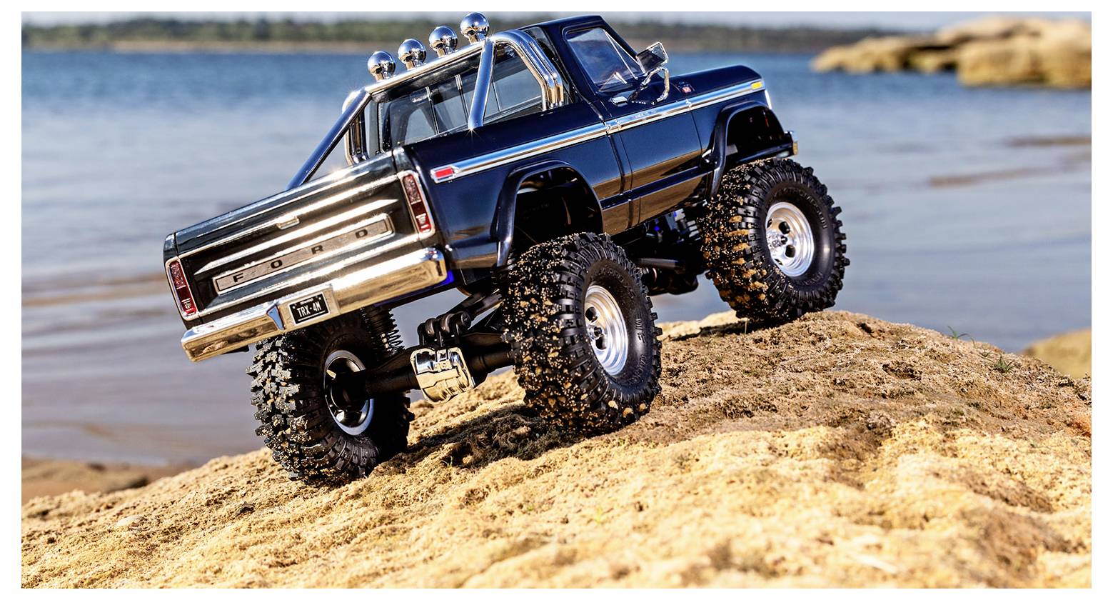 A blue remote-controlled off-road truck with large tires climbs a sandy hill by a lake, showcasing its rugged design and capabilities.