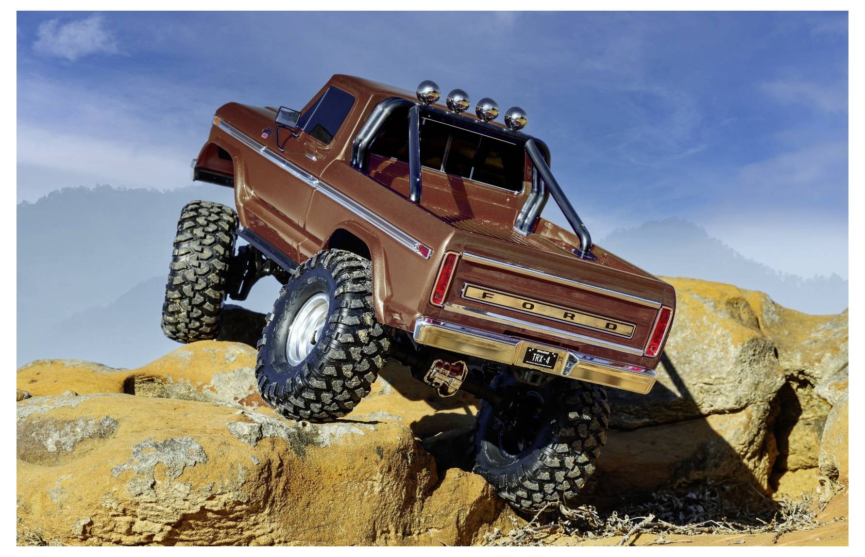 A rugged toy truck with large tires navigates over rocky terrain under a clear sky, showcasing off-road capabilities.