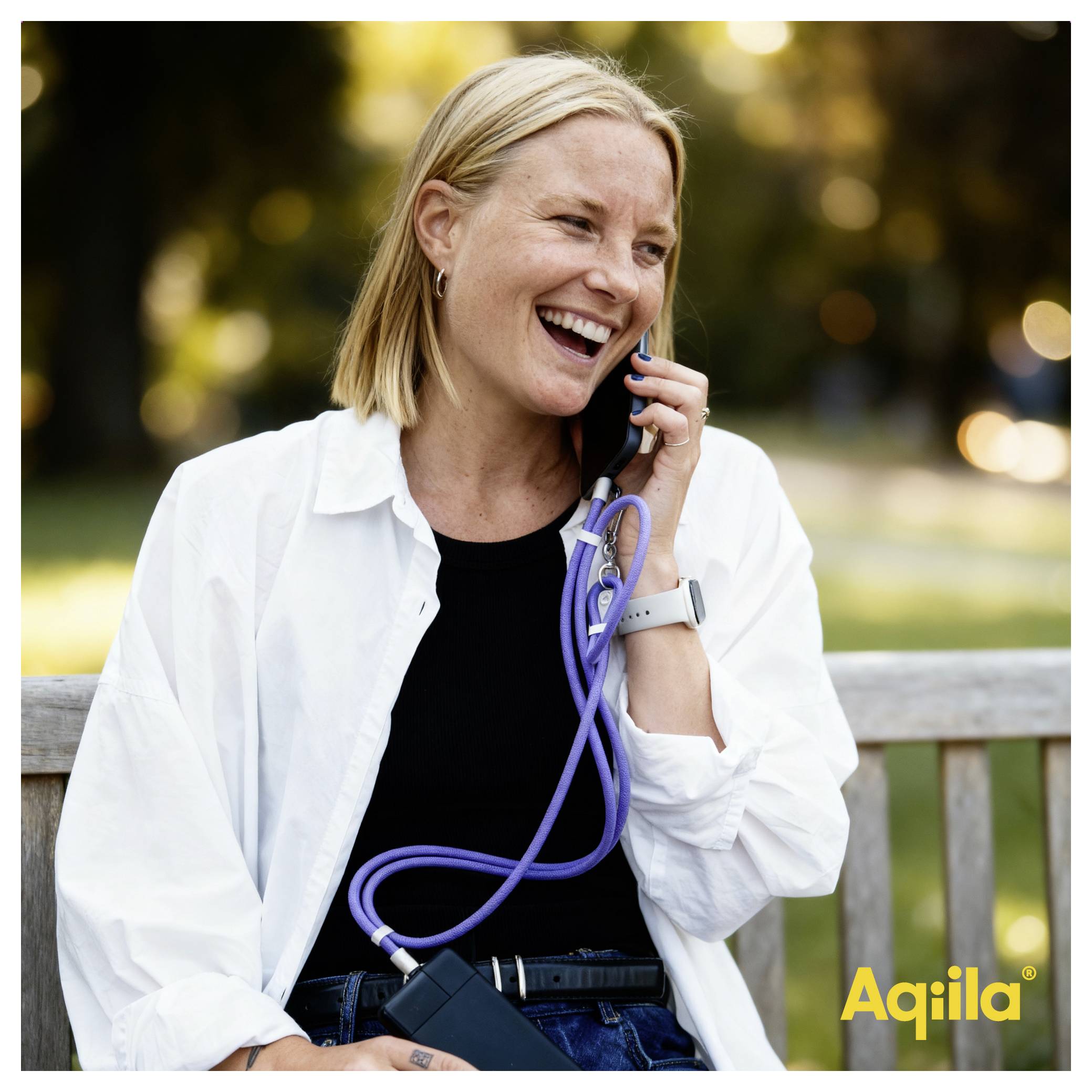 A person smiling while talking on a phone, sitting on a bench outdoors. They have a relaxed posture and appear cheerful.