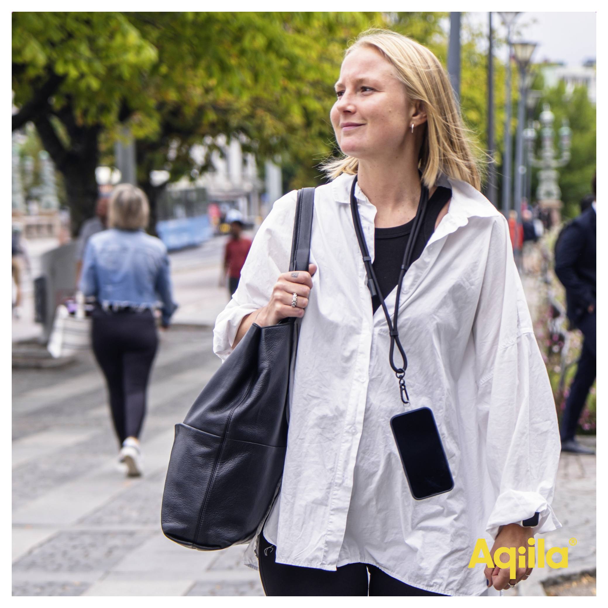 A person in a white shirt and black shoulder bag walks along a city street, appearing content. Other pedestrians and greenery are visible.