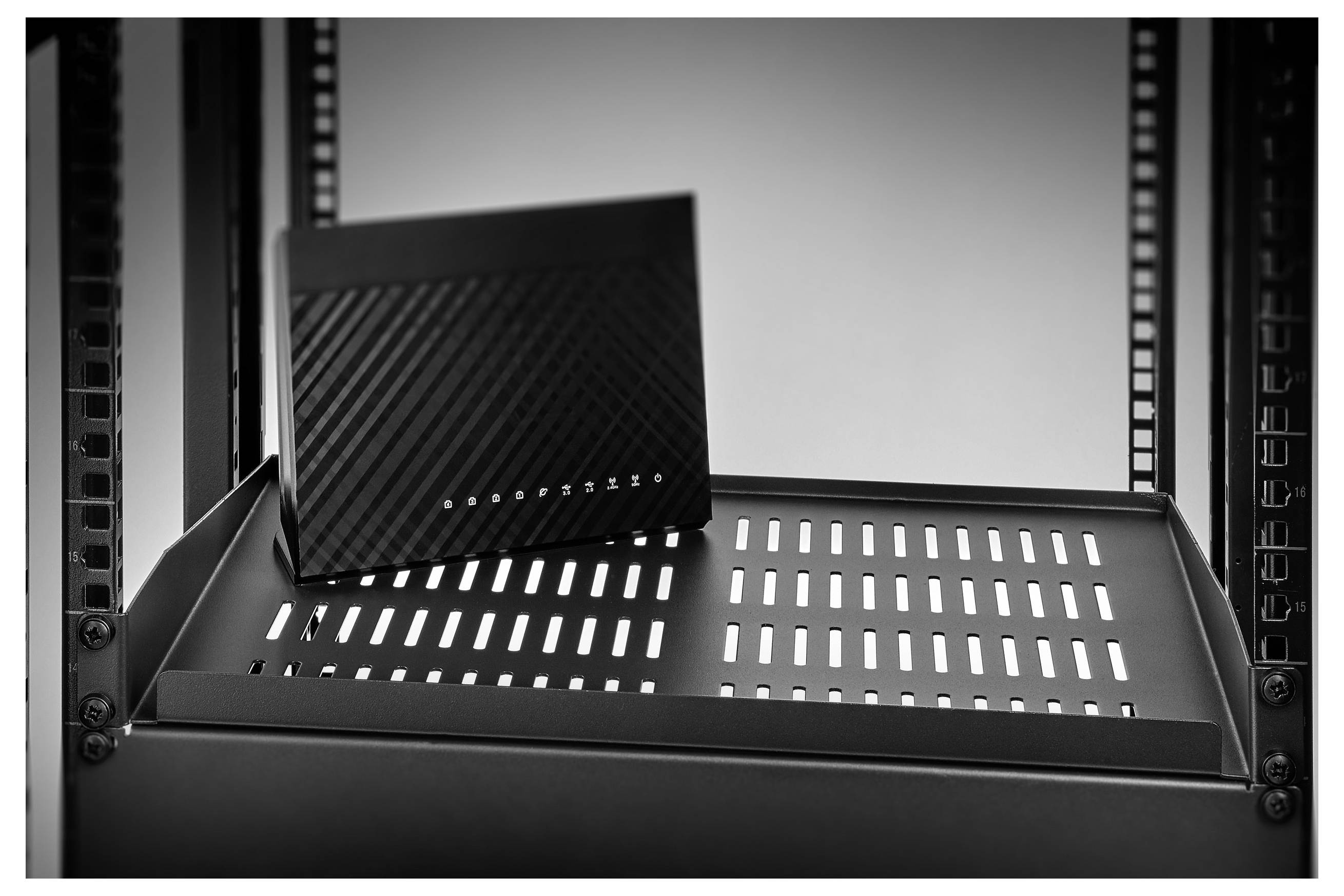 Black network router or modem on a server rack shelf with front indicator lights and diagonal texture.<br>