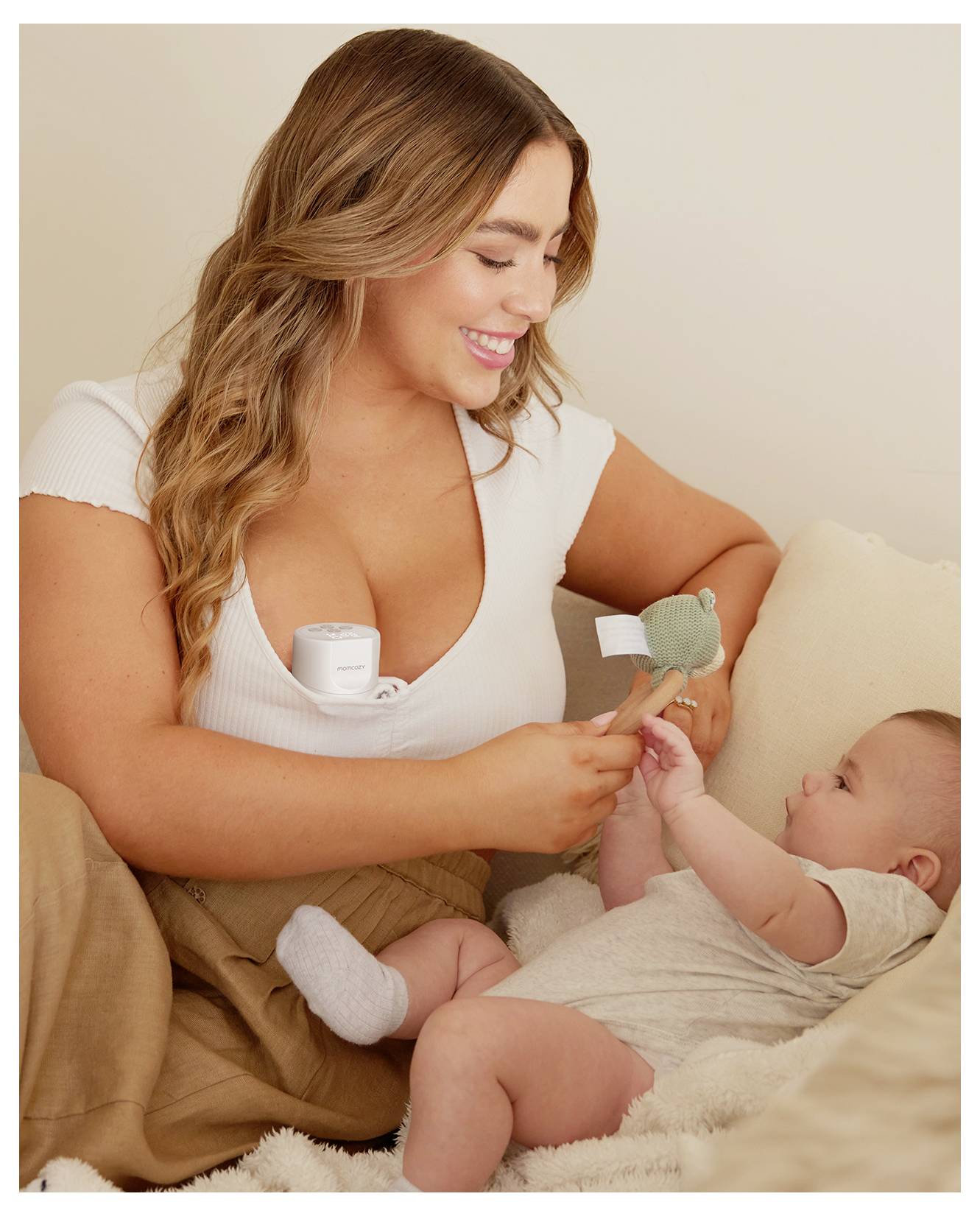 A woman with long hair holds a small toy, smiling at an infant lying beside her on a beige couch. The infant reaches for the toy.