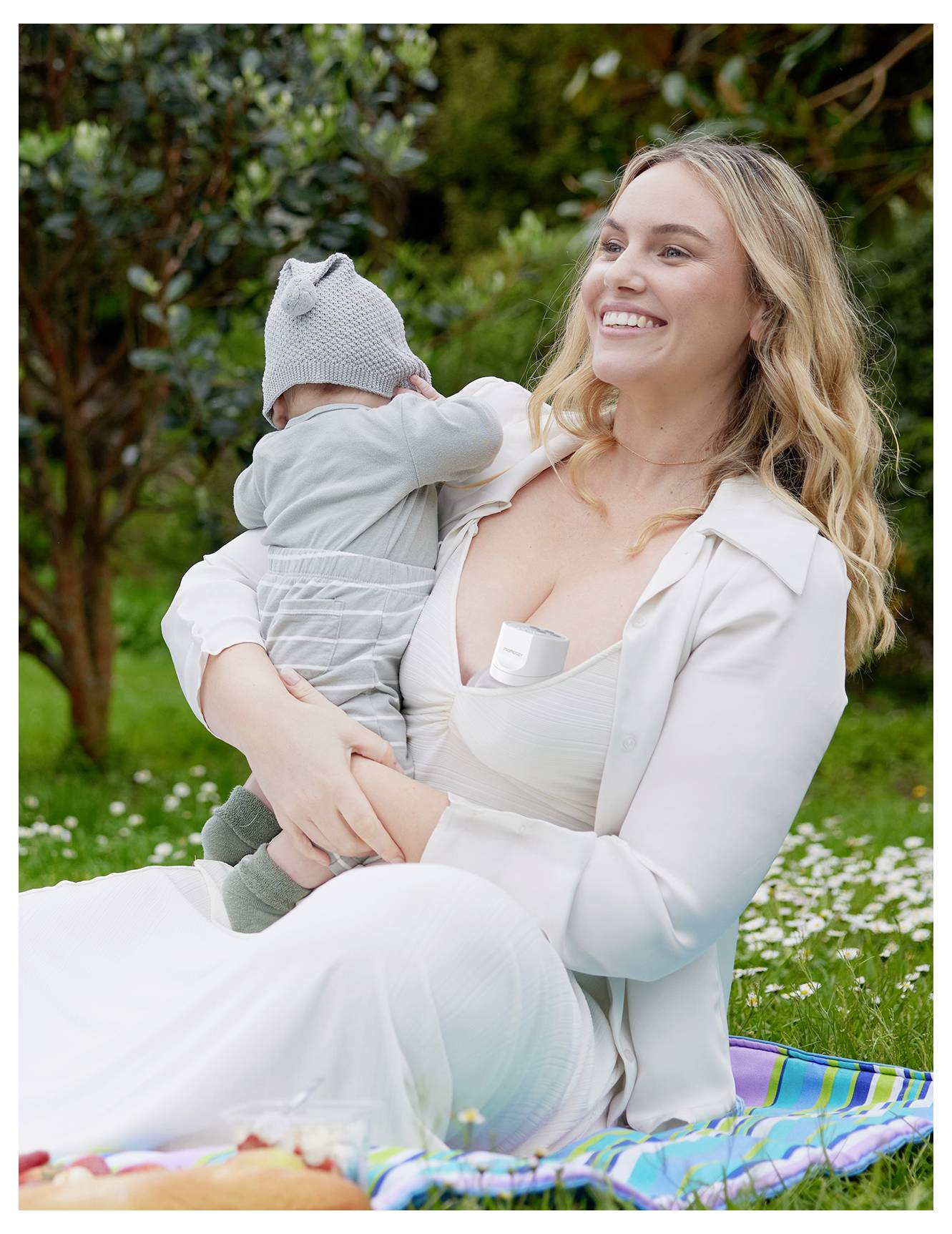 A mother holds her baby in a park setting, both dressed in light-colored clothing. They are sitting on a striped picnic blanket with greenery around.