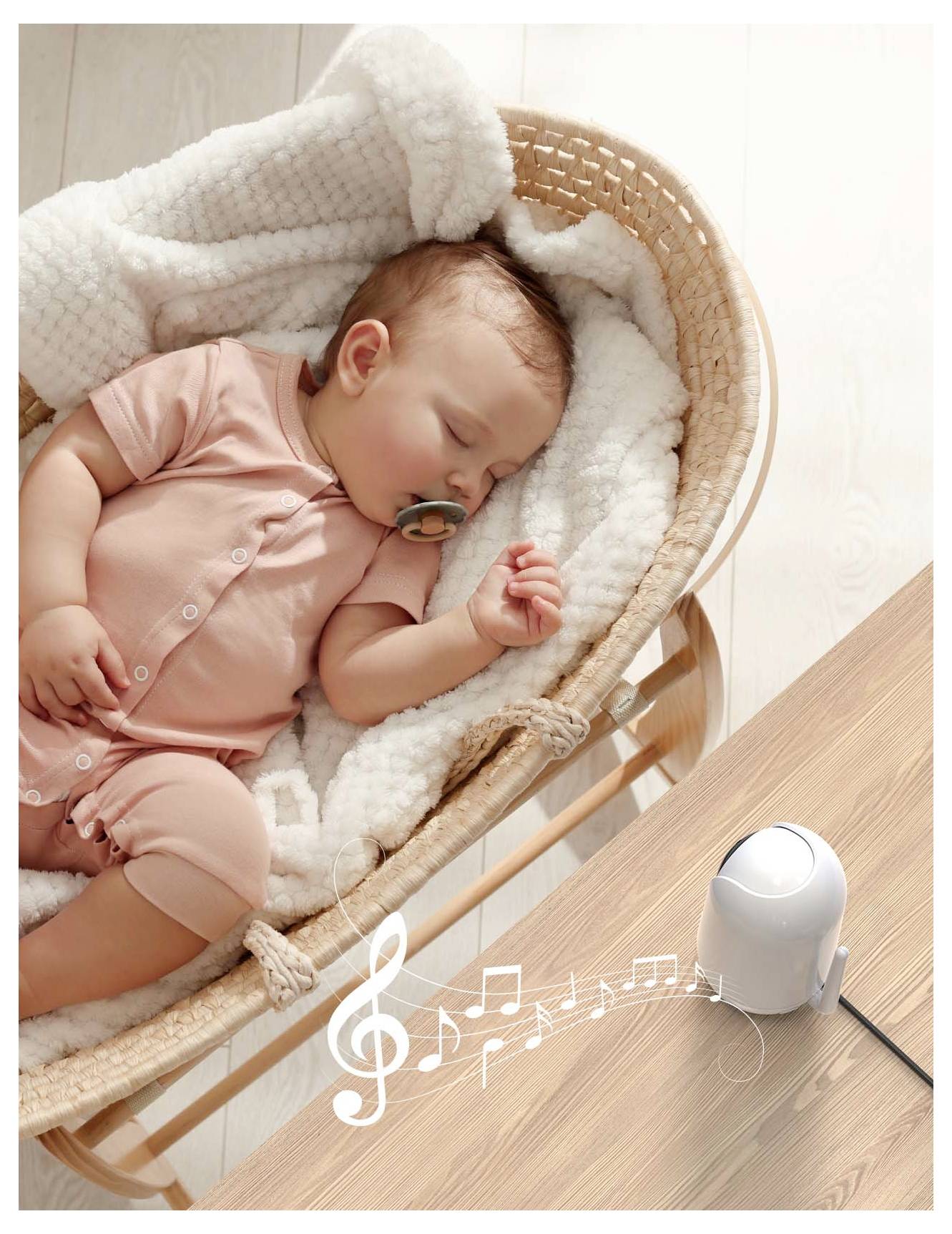 A baby sleeps in a wicker basket with a monitor playing music notes nearby.