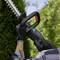 A person uses a cordless hedge trimmer to shape a bush in a garden. The focus is on the tool and the bush, highlighting its ergonomic design.