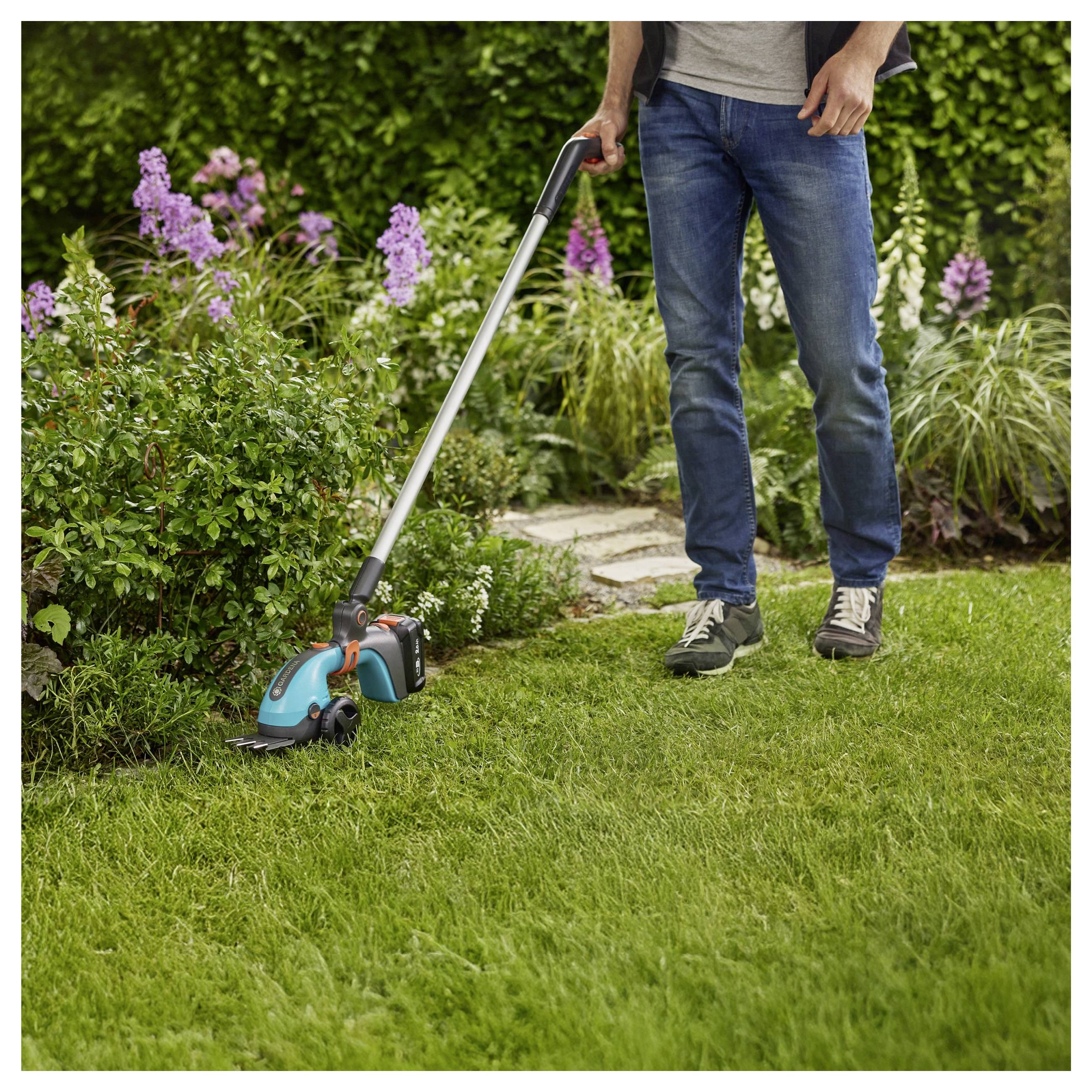 A person is using a grass trimmer to cut the edge of a garden lawn. The garden has green grass, a stone path, and flowering plants.