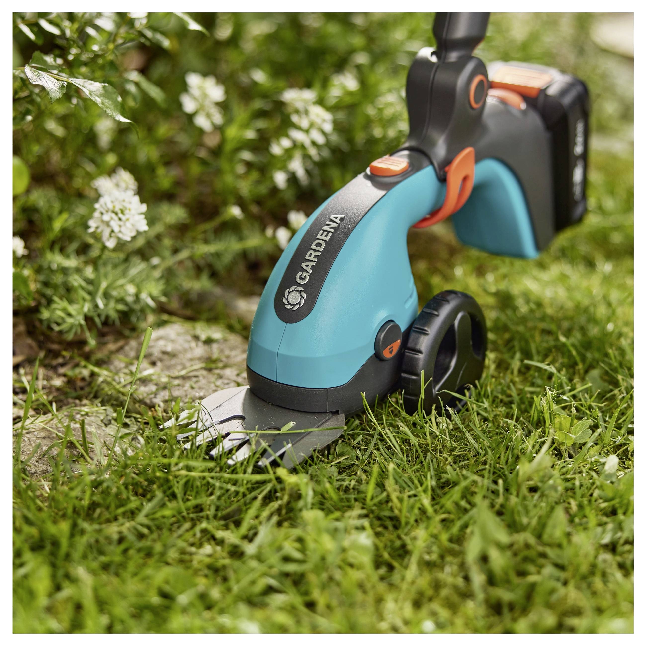 'A Gardena cordless grass shear trimming grass edges near small white flowers in a garden setting.'