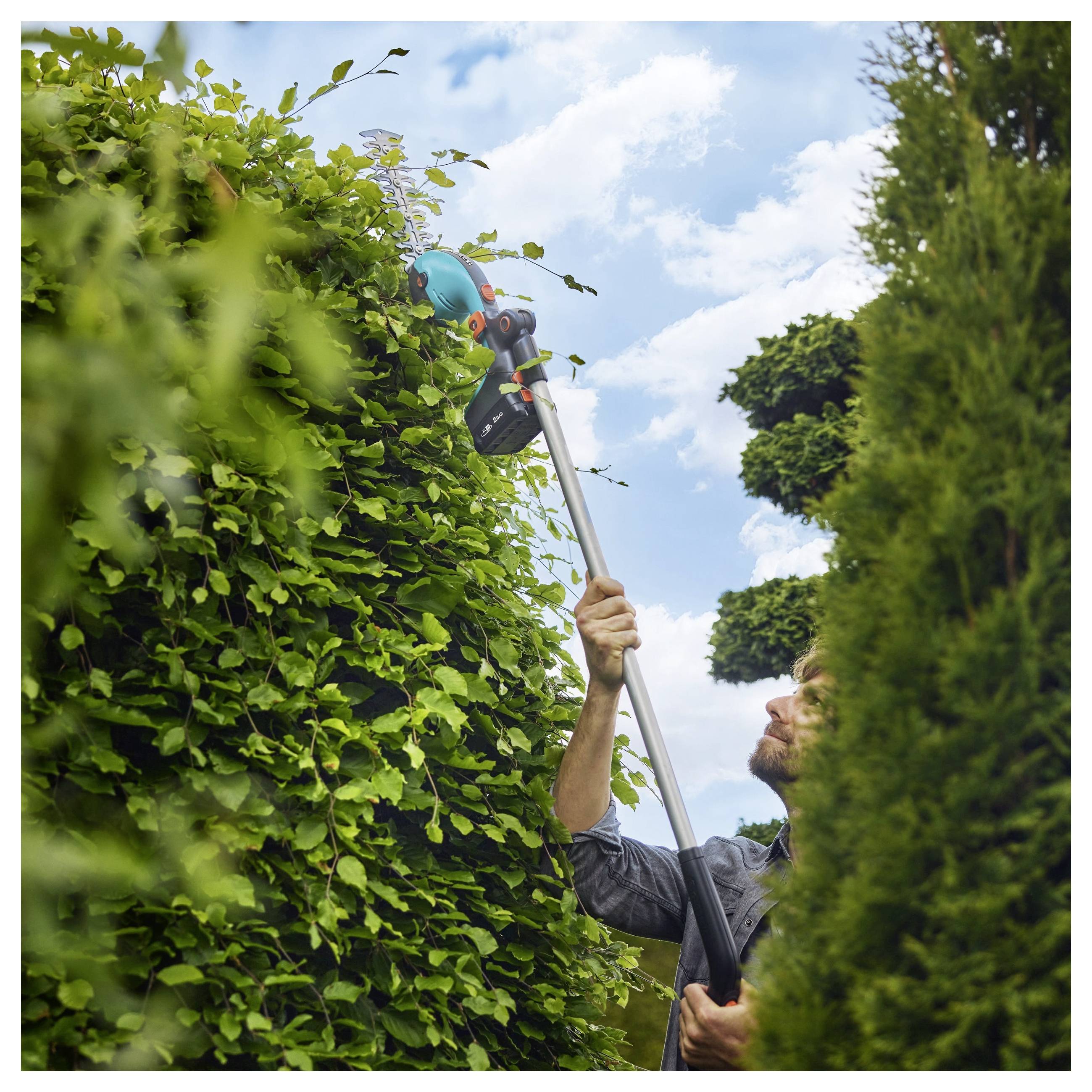 Man trimming tall hedges with a long-handled electric trimmer against a backdrop of a cloudy sky.