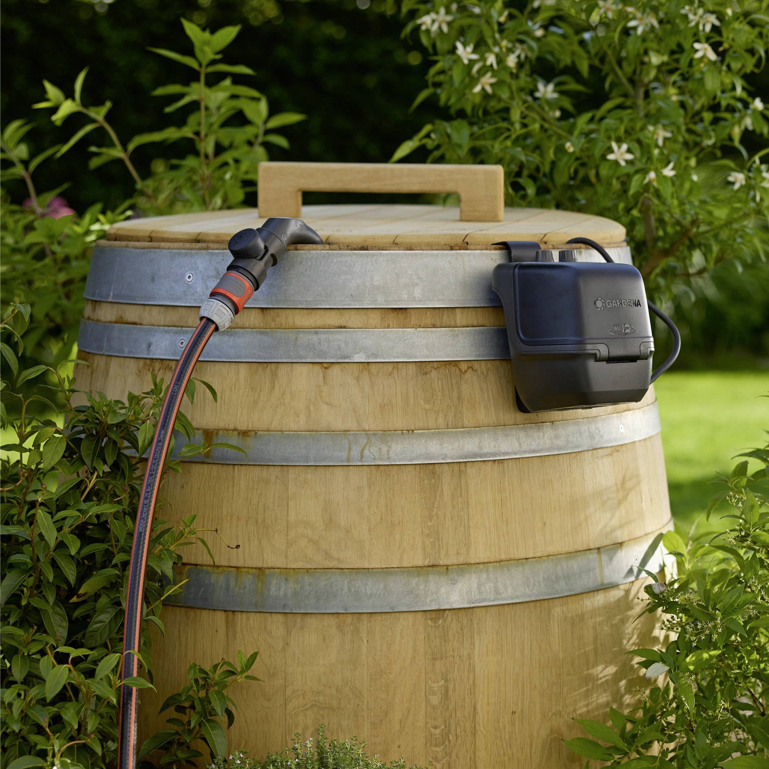 A wooden rain barrel with a water-saving valve attached, placed in a garden setting with green plants, under clear daylight.