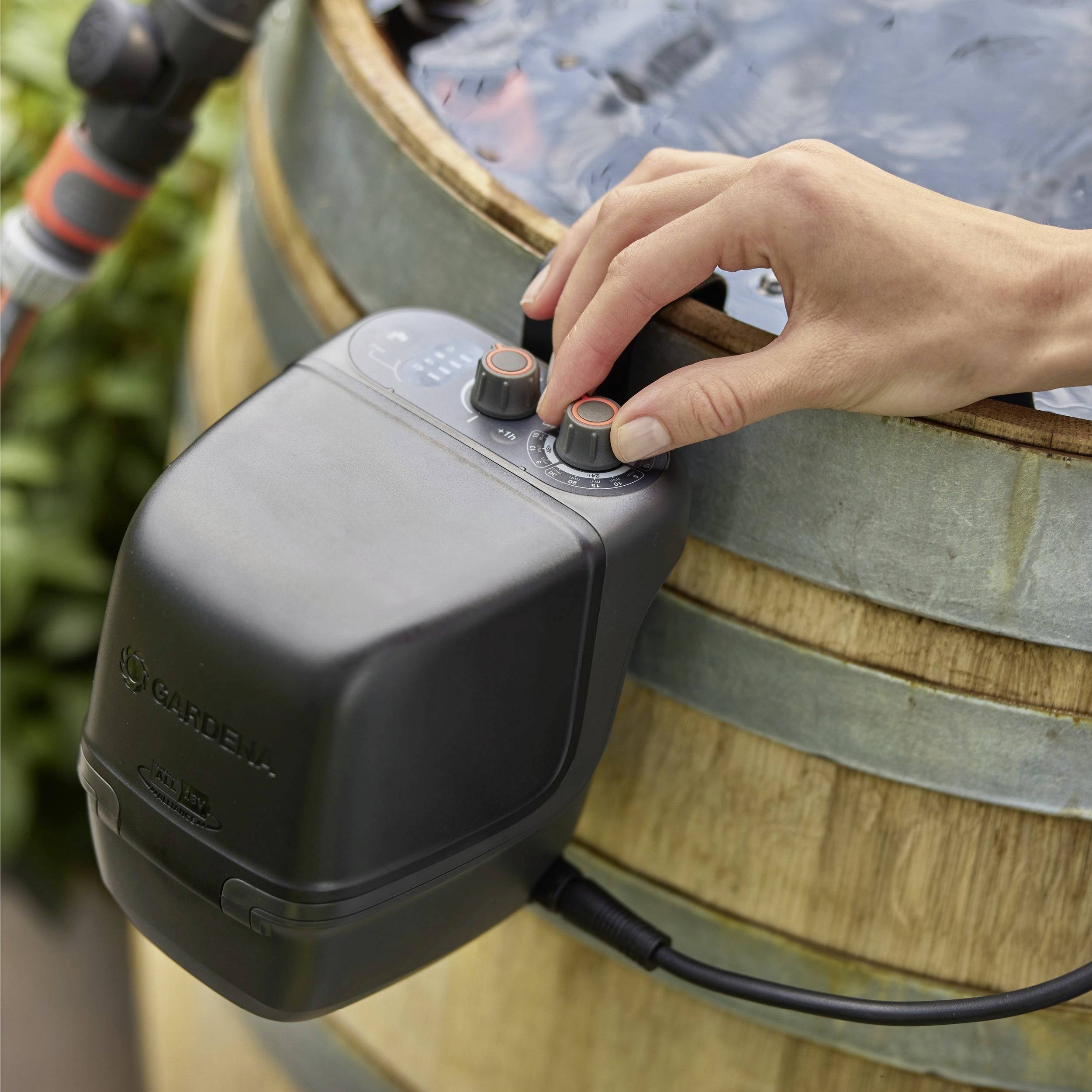 A hand adjusts a dial on a water timer connected to a garden barrel, controlling water flow for irrigation.