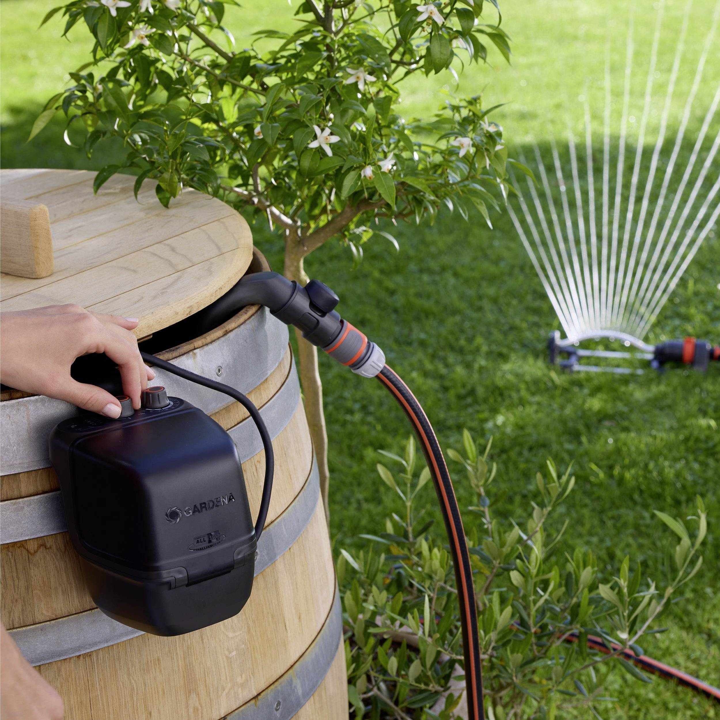 A person installs a Gardena water timer on a rain barrel in a garden. A sprinkler waters the lawn nearby.
