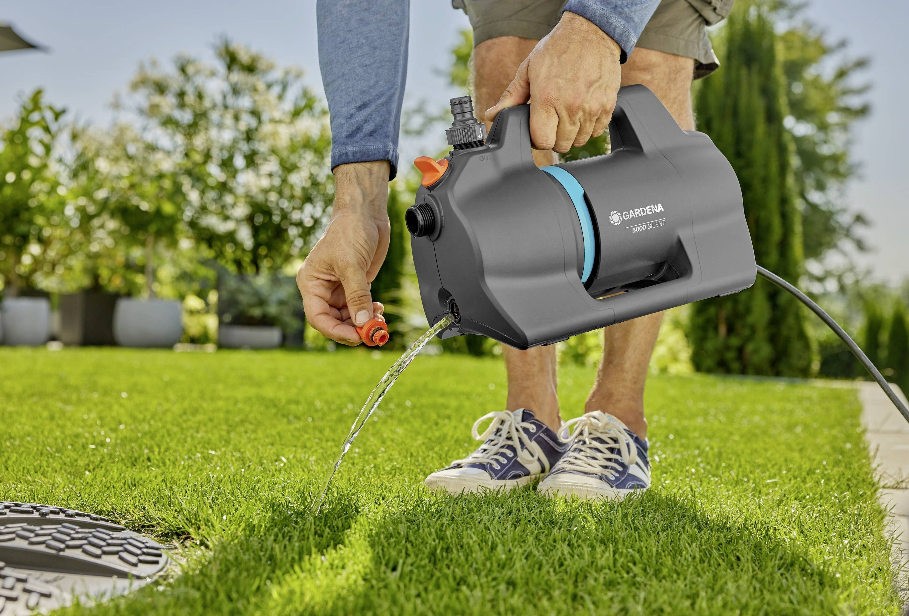 A person waters a lawn using a handheld Gardena garden pump, with water streaming onto the grass. Sunlit background with plants.