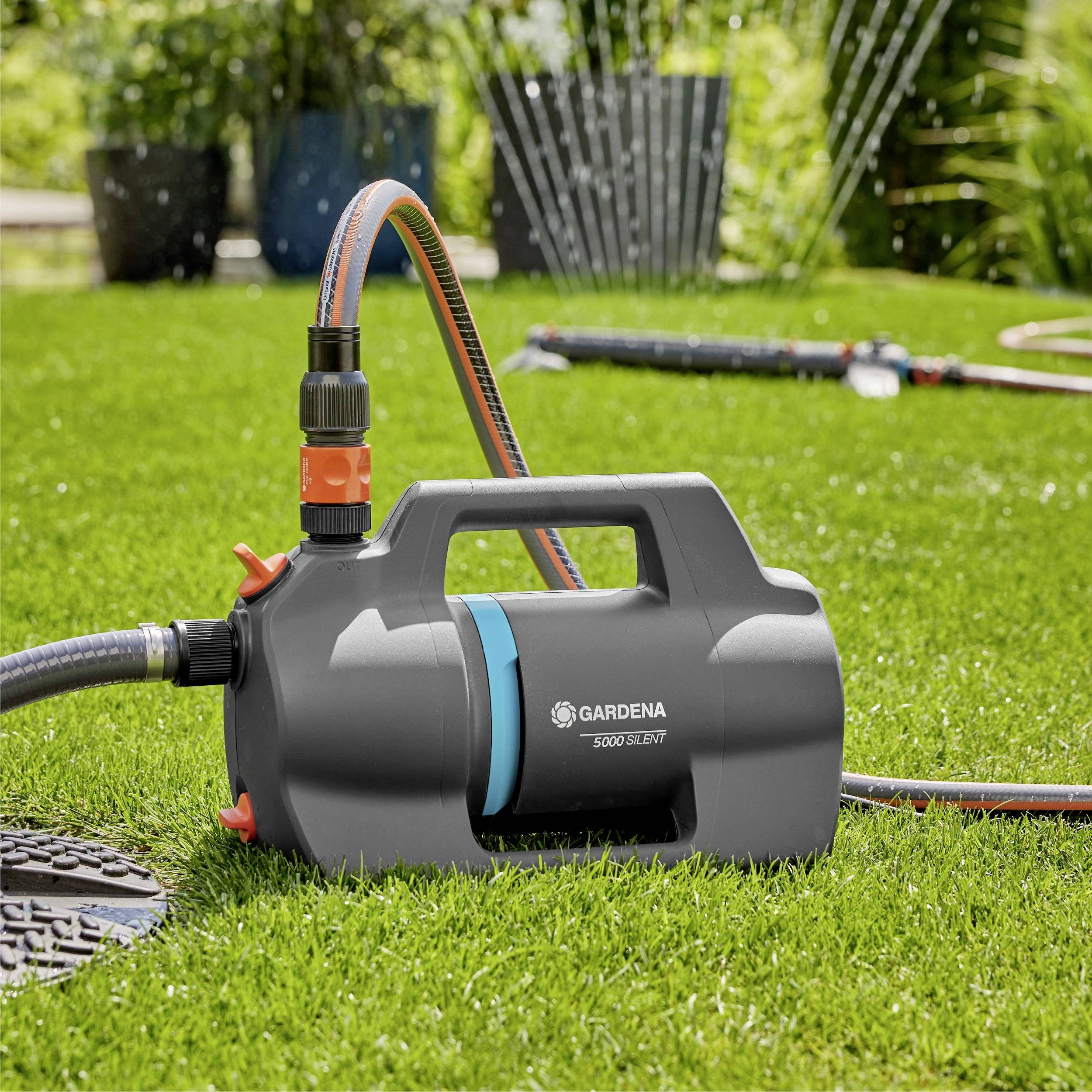 A portable garden pump on grass with sprinklers in the background. The pump is branded 'Gardena 5000/5E'.