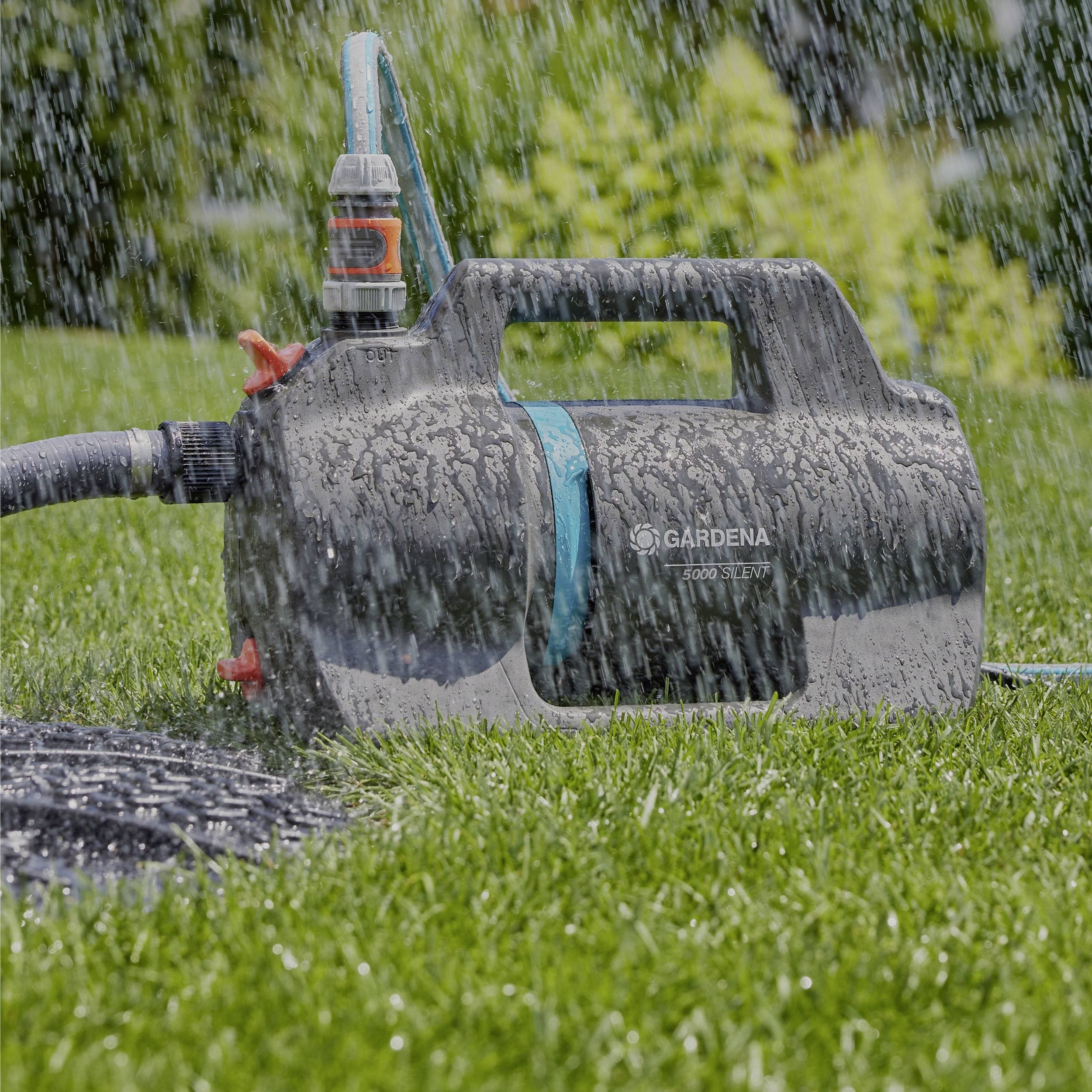 A black and blue electric garden water pump sprays water over a grassy lawn, with trees blurred in the background.