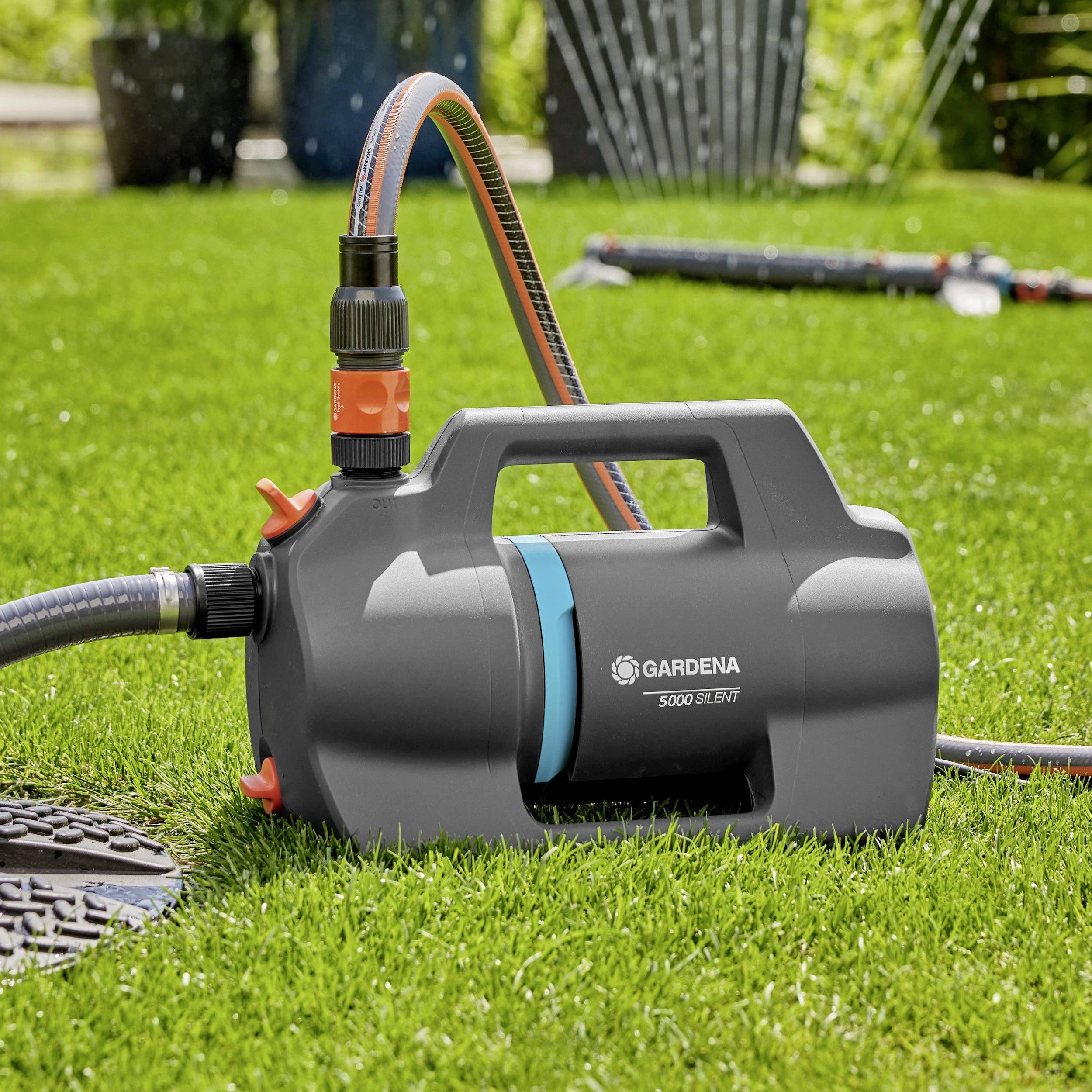 A Gardena 5000 Silent water pump on grass, connected to a hose, with garden in the background, indicating its use for irrigation.