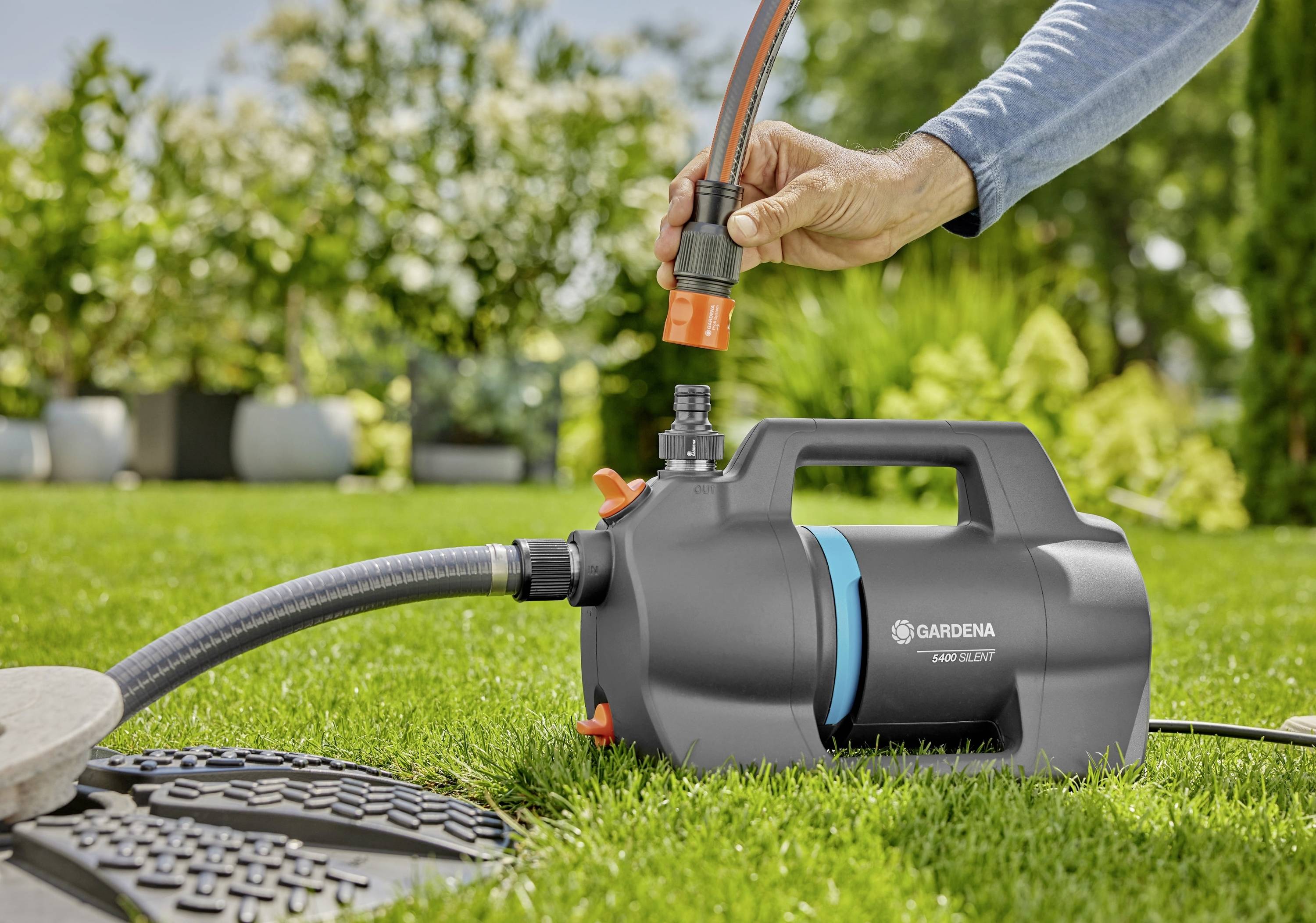 A person is connecting a garden hose to a Gardena water pump on a lawn, surrounded by plants.