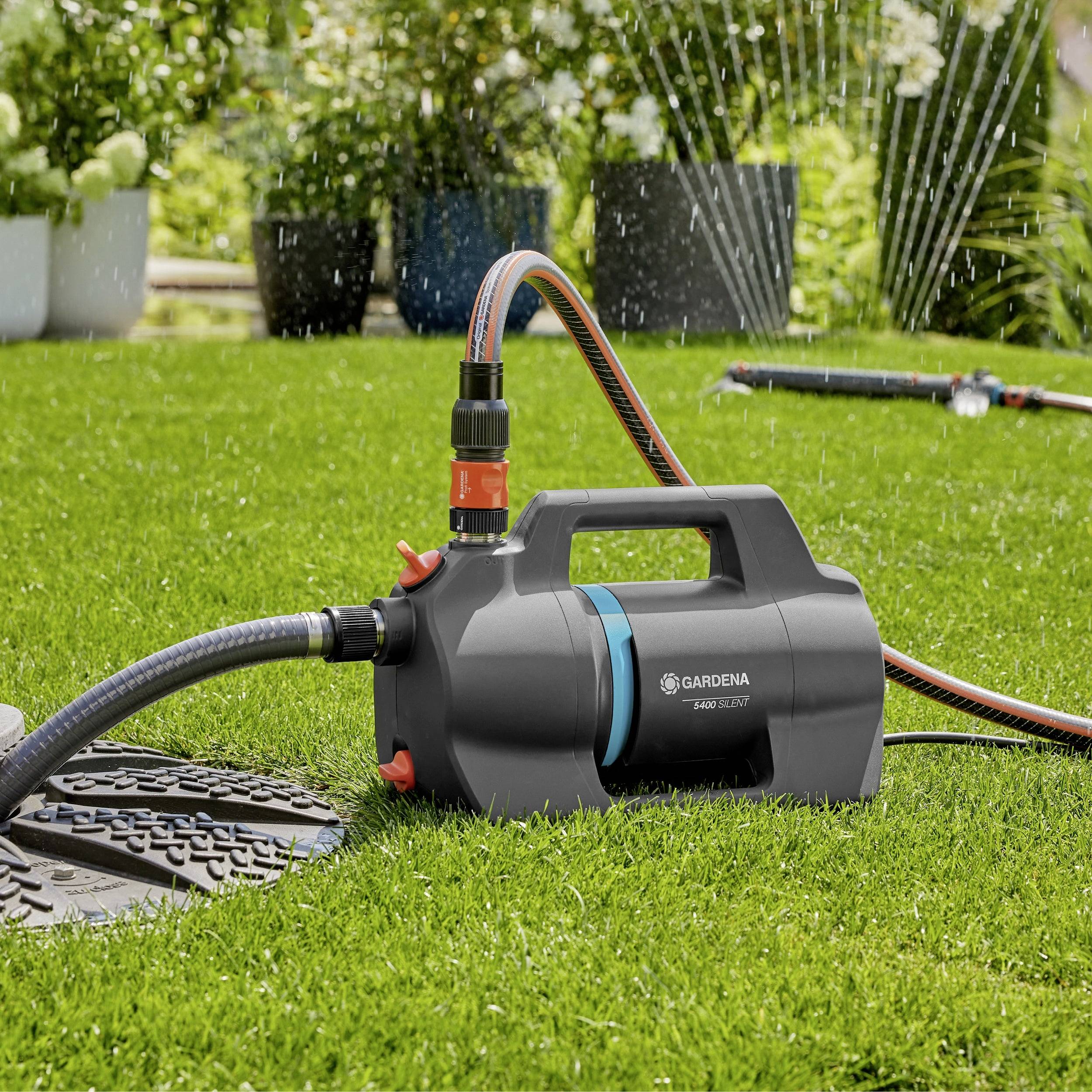 A garden pump sits on green grass with hoses attached, actively watering the lawn and nearby plants.