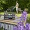A garden water pump in use, surrounded by flowers and greenery, displaying the brand name. It's raining, emphasizing the pump's utility.