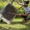 A person smiles while washing a wheelbarrow with a garden hose in a sunny garden, surrounded by greenery.
