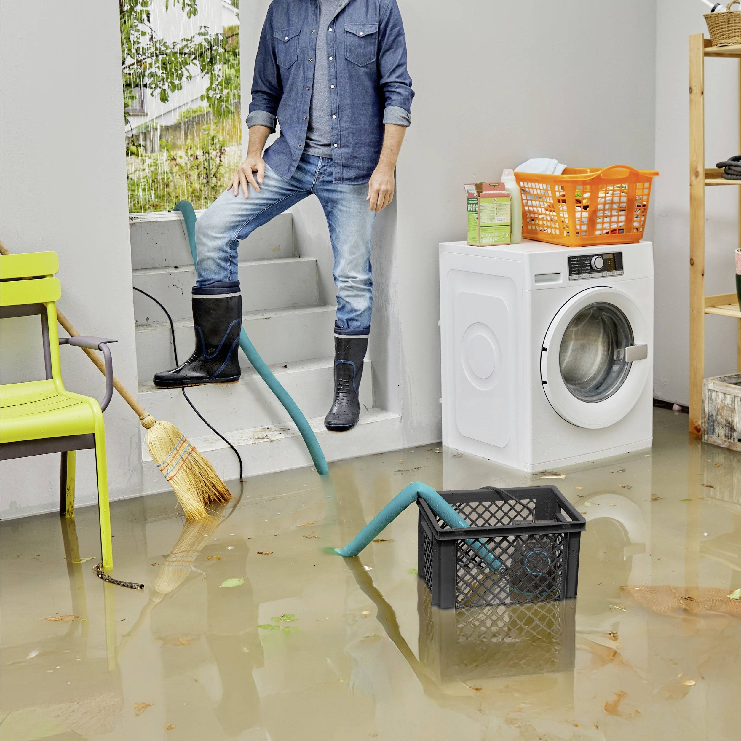 A person in boots stands in a flooded laundry room with a washing machine, chair, broom, and basket partially submerged in water.