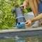 A person adjusts a garden watering device on a stone ledge near ferns, ensuring proper irrigation setup in a garden setting.