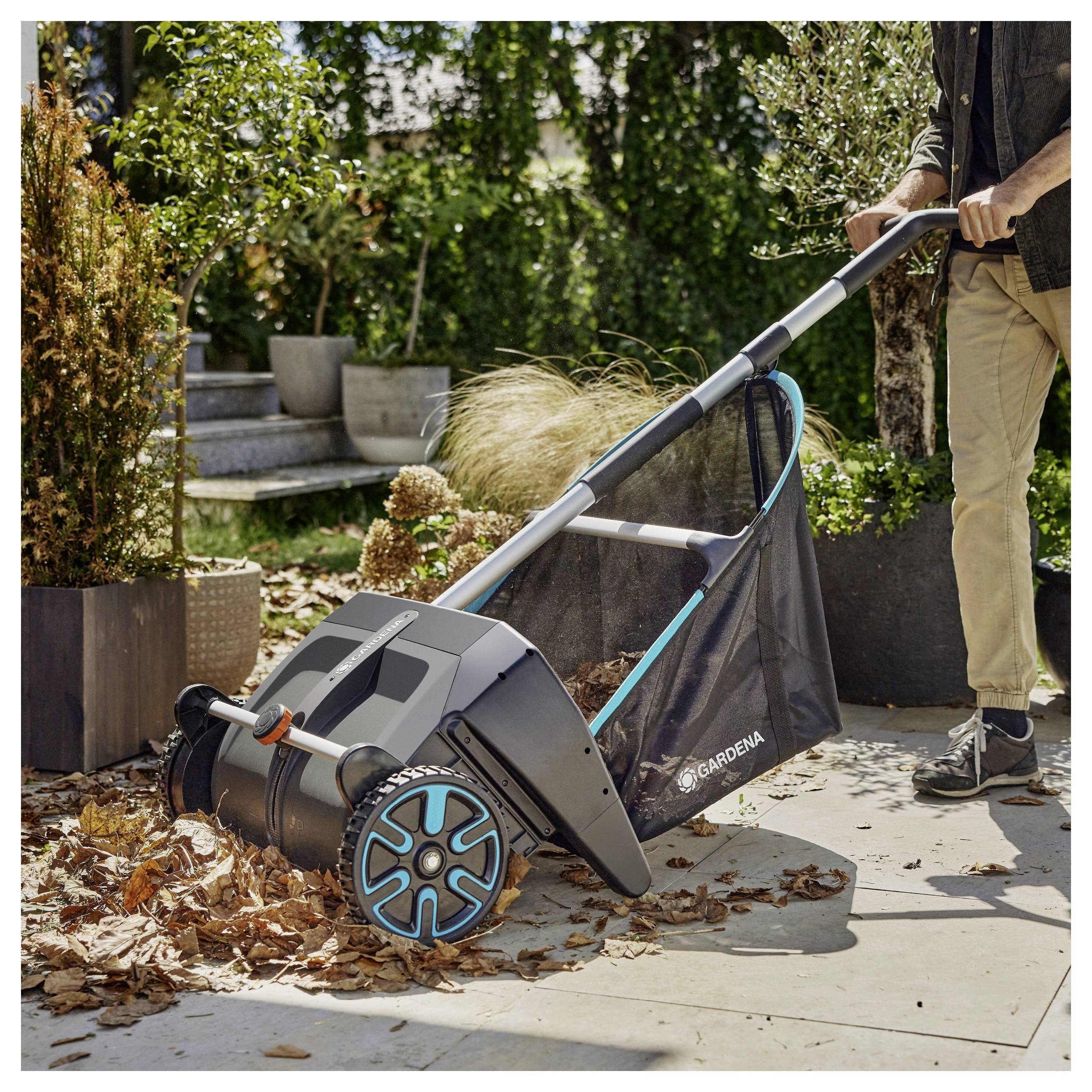 A person uses a garden leaf collector on a patio, gathering fallen leaves. The tool is wheeled, with a mesh bag for leaf collection.