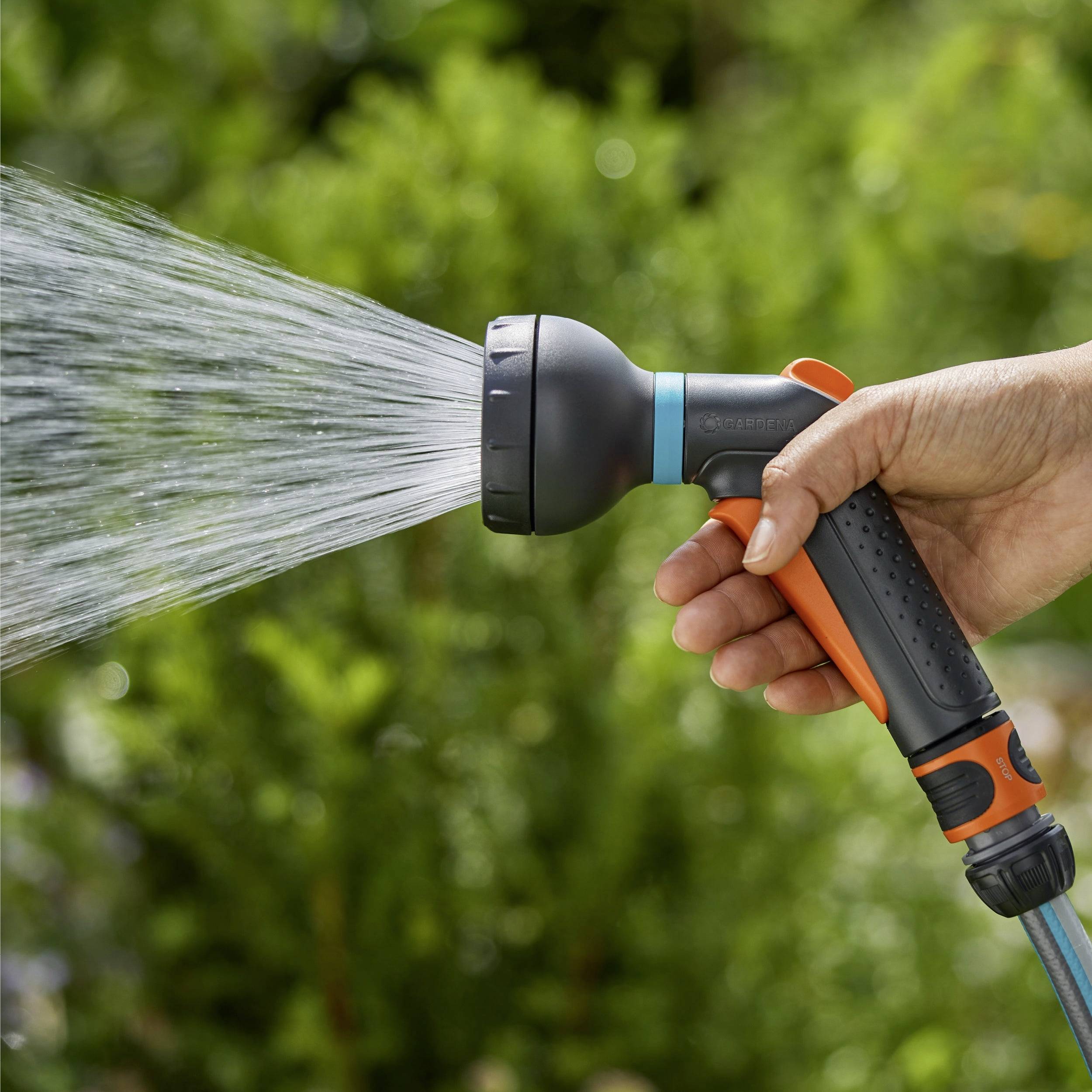 A hand holds a garden hose with a spray nozzle, watering green plants in a garden setting.