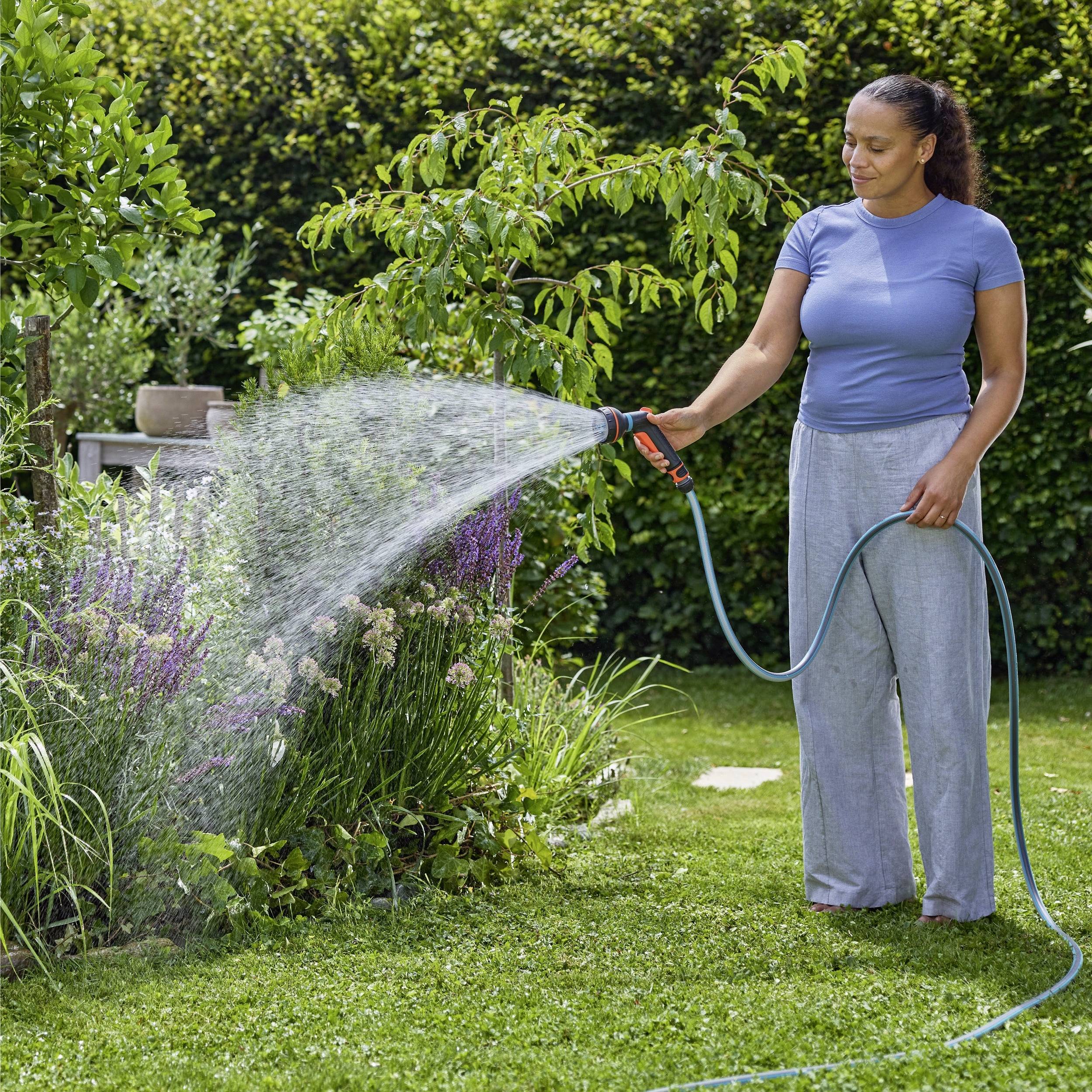 A person in a garden uses a hose to water plants, surrounded by lush greenery and flowers, on a sunny day.