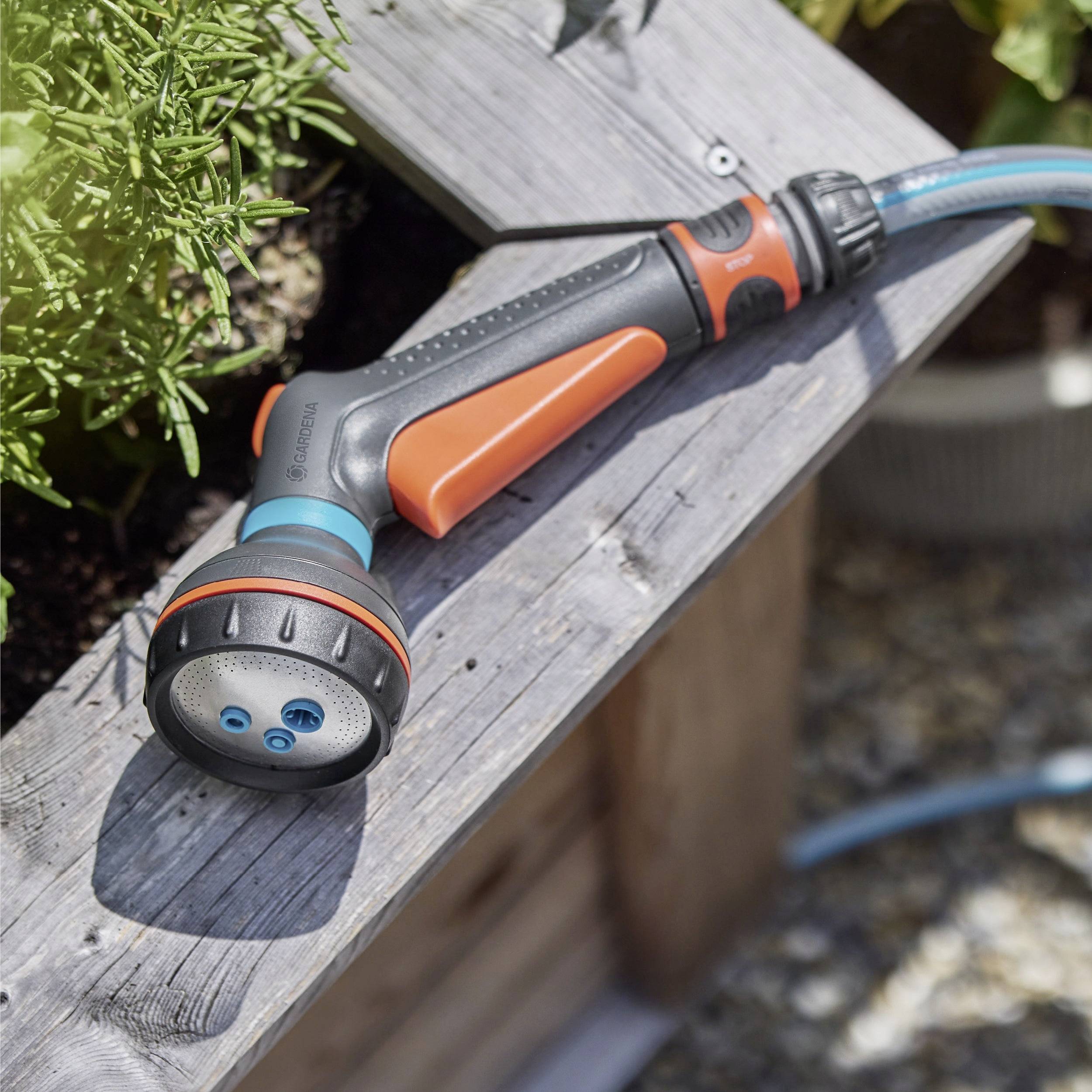 A garden hose nozzle with orange and black grips lies on a wooden bench, surrounded by green plants.