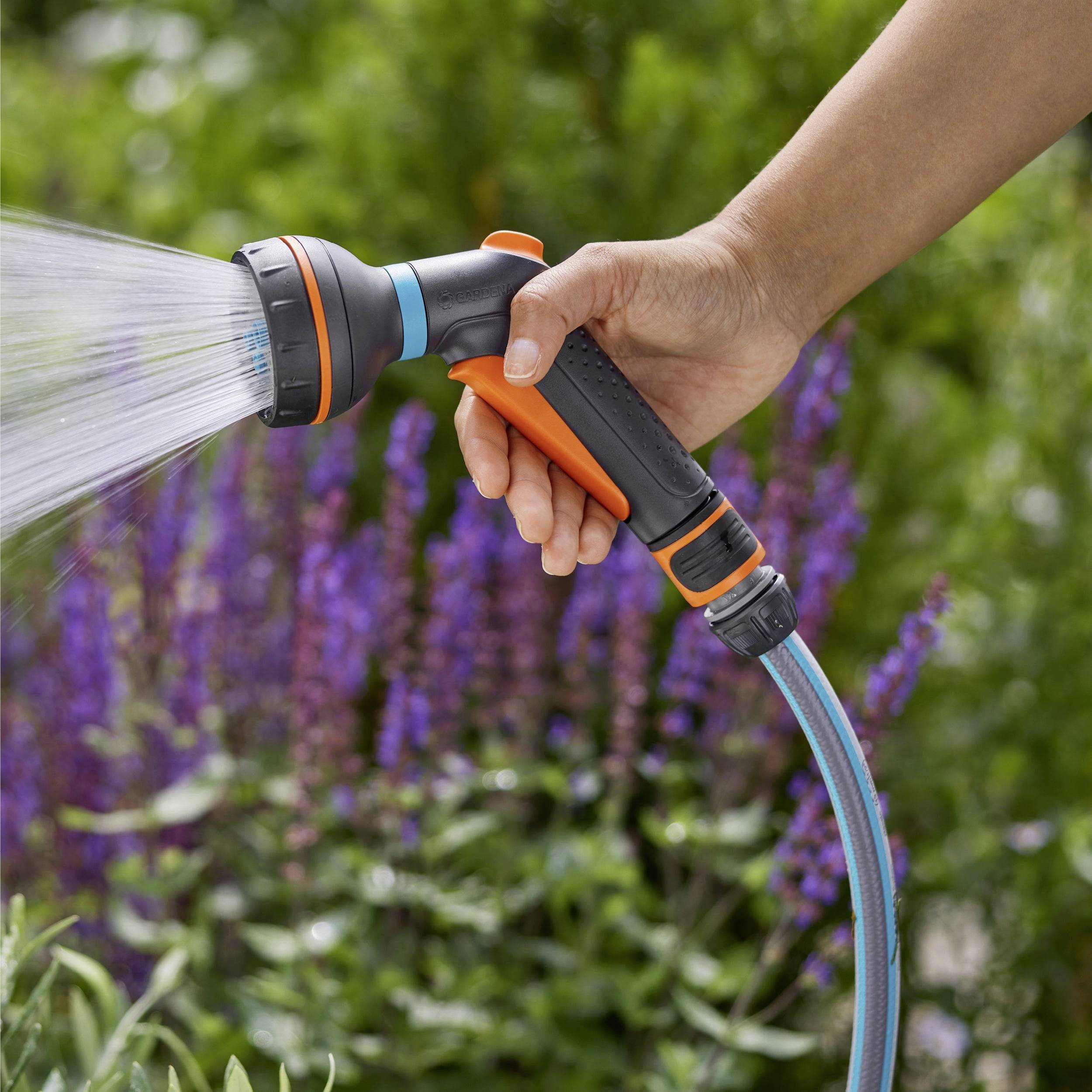 A hand holding a garden hose with a spray nozzle, watering purple flowers in a garden setting.
