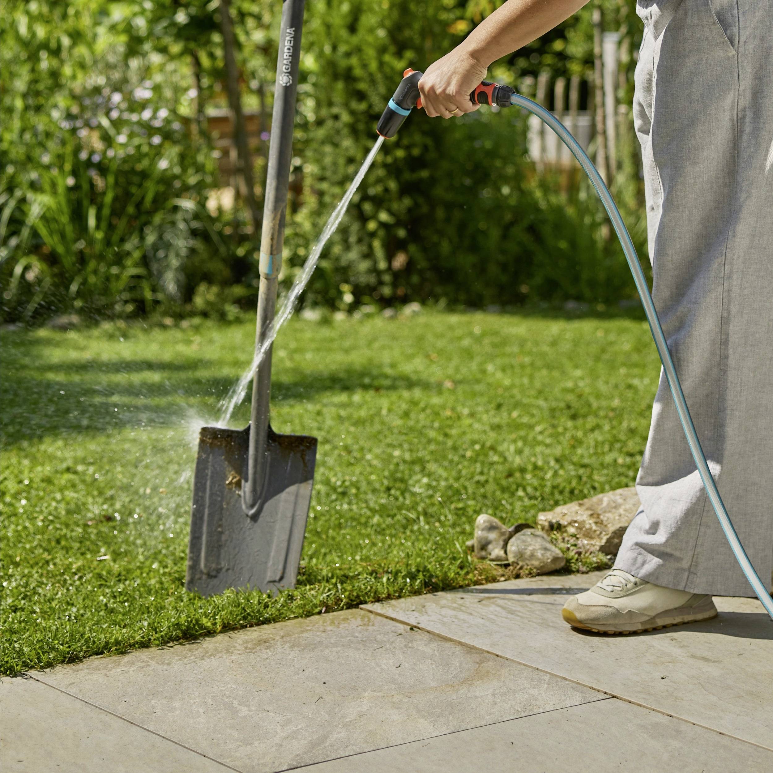 A person is watering a garden with a hose on a sunny day, next to a shovel standing upright in the grass.