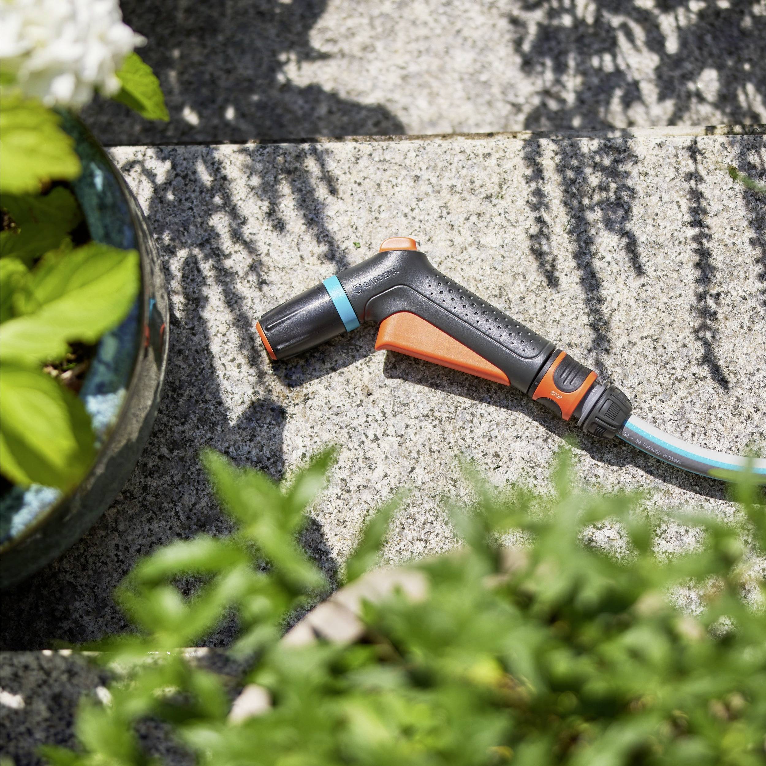 A gardening hose nozzle, with an orange and black design, lies on a stone surface next to potted plants, ready for watering.