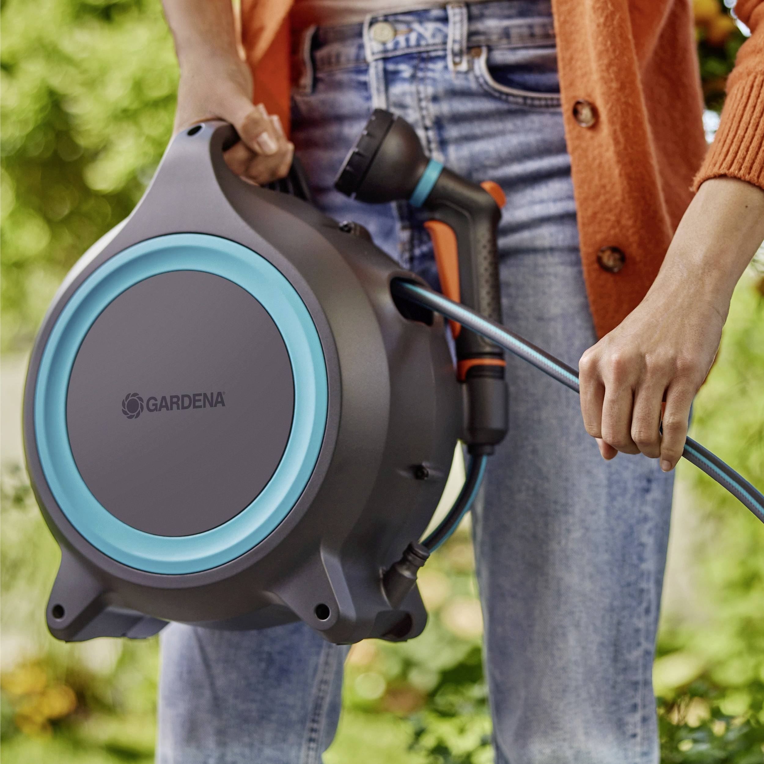 Person holding a gray and blue GARDENA hose reel in a garden, wearing an orange sweater and jeans.