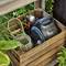 A garden hose cart and nozzle are neatly stored in a wooden crate, surrounded by lush green leaves in a garden setting.