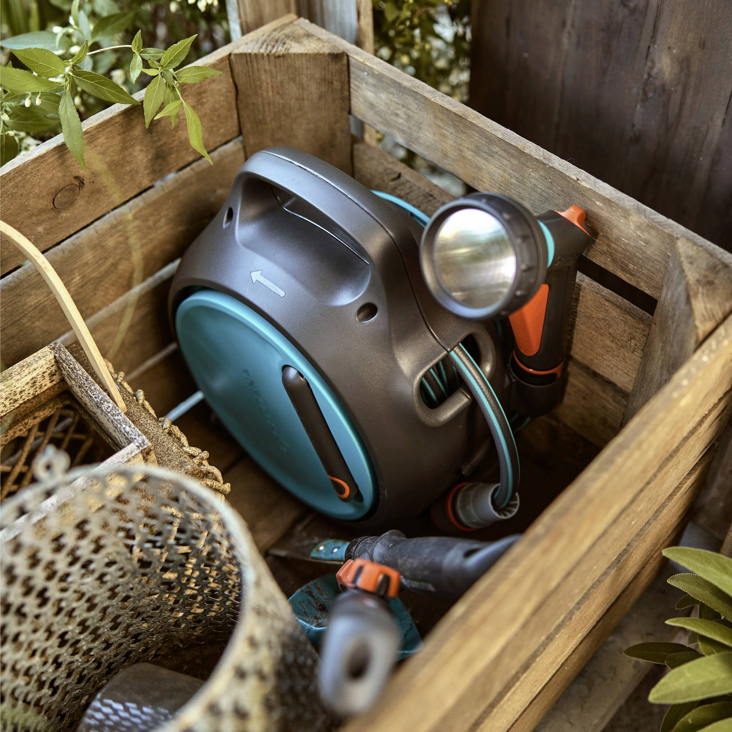 Garden hose reel with attached sprayer stored in a wooden crate, surrounded by gardening tools and plants, suggesting a gardening setup.
