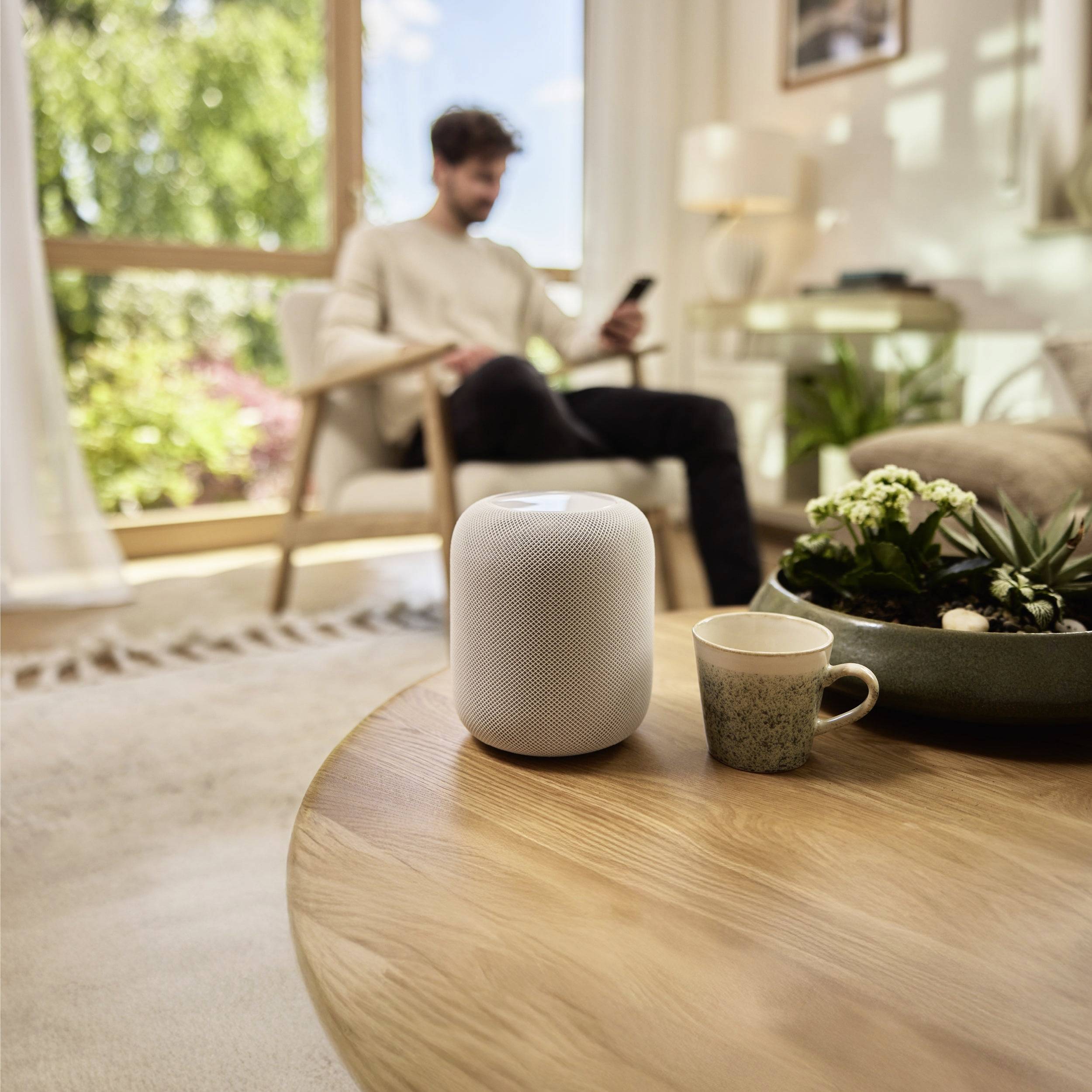 A smart speaker and a coffee cup on a wooden table, with a person using a smartphone in the background, seated in a cozy room.