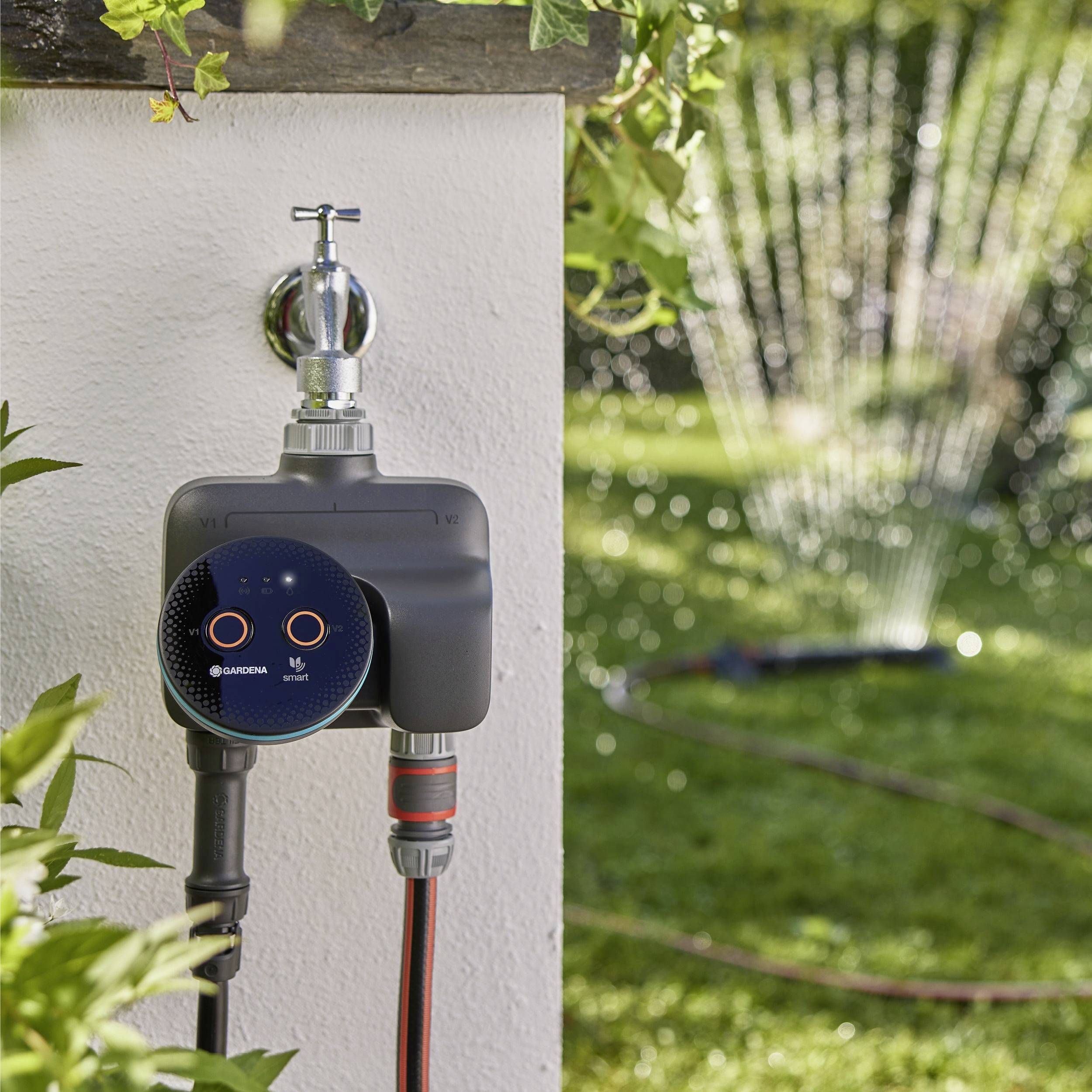 A digital water timer is attached to an outdoor faucet, controlling a hose that waters a garden with an oscillating sprinkler.