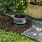A robotic garden tool is watering plants near a potted shrub on a stone pathway. The device is small, round, and black with a water hose attached.