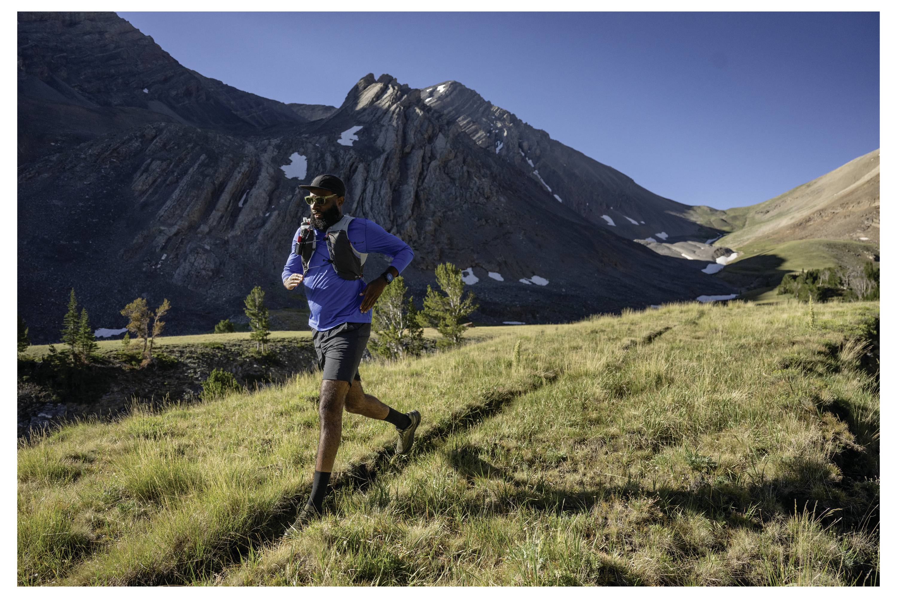 A person is running on a grassy hillside with mountains in the background, wearing a blue top and carrying a hydration pack.