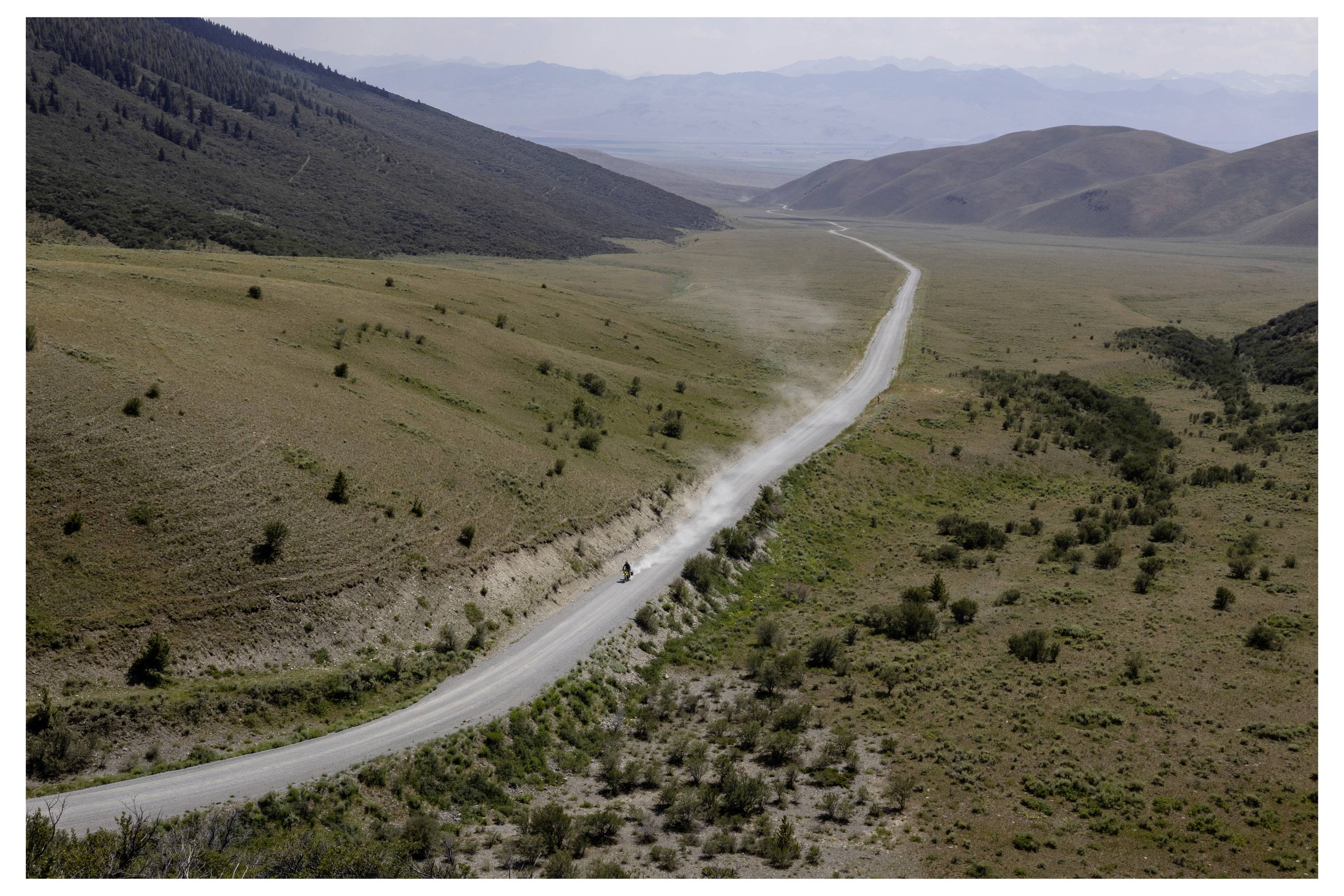 A car drives along a long, winding dirt road through a vast, mountainous landscape under a clear sky.