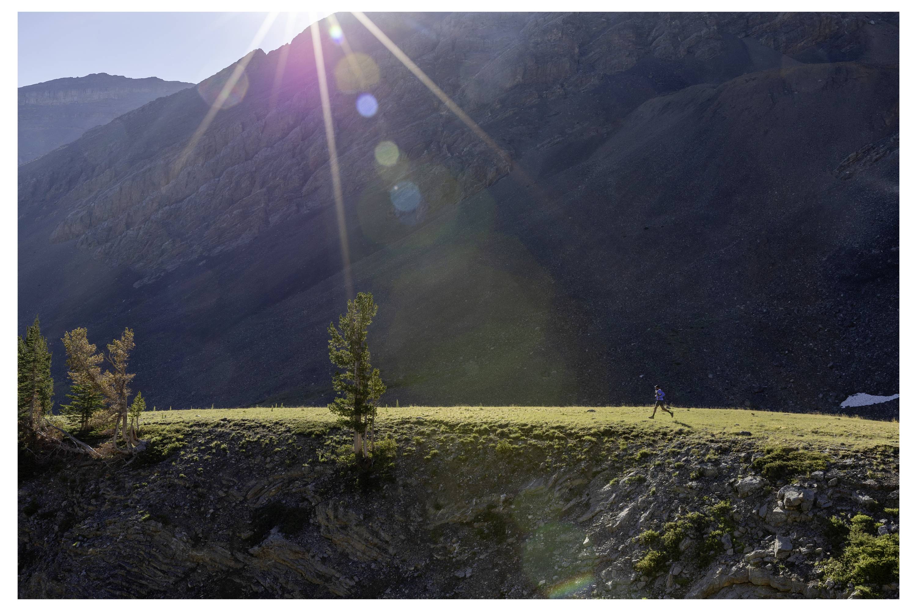 A person walks on a grassy ridge under a bright sunbeam with a mountainous backdrop. Trees are scattered along the ridge.