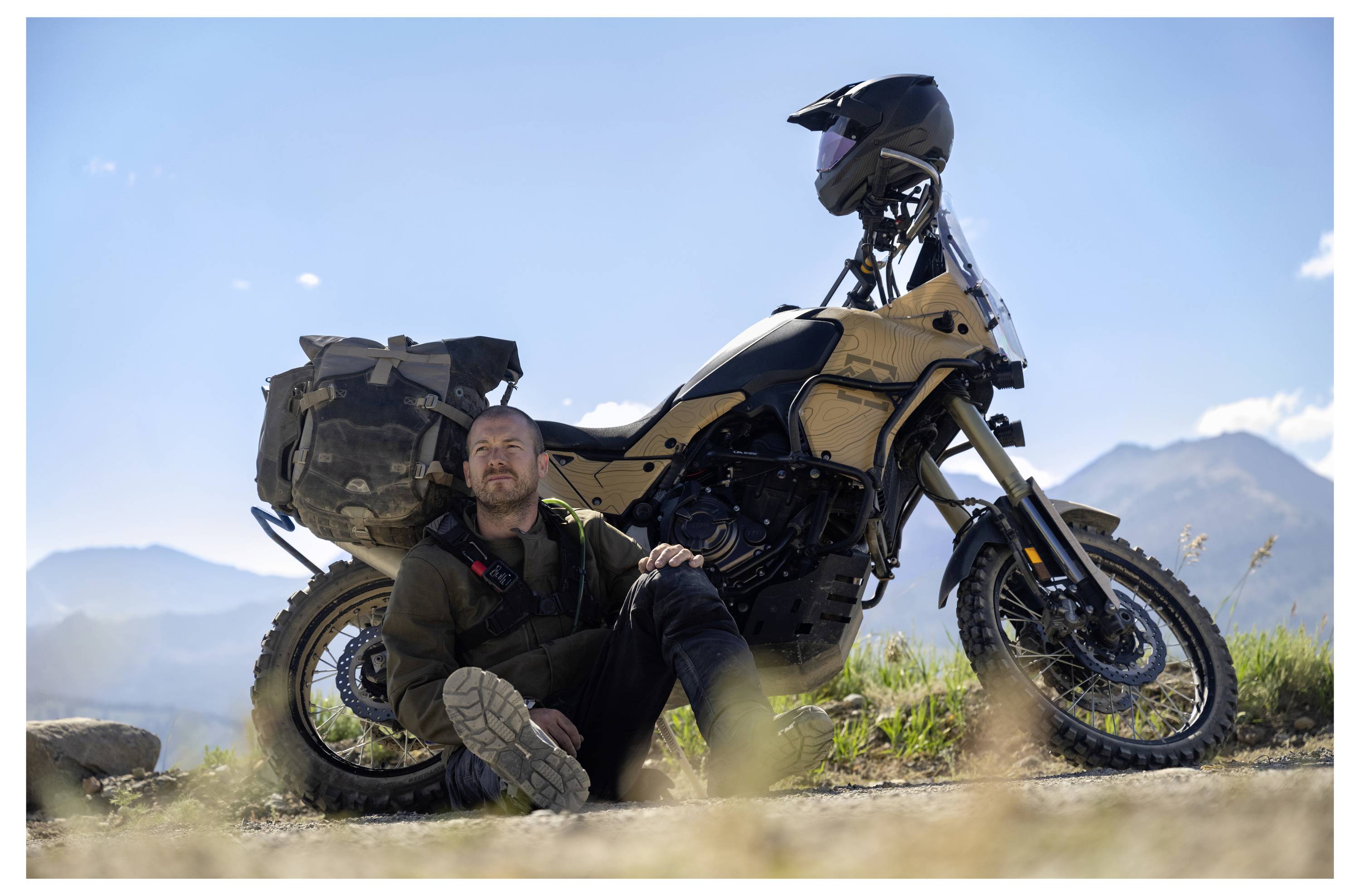 A person sits on the ground, leaning against an adventure motorcycle in a scenic, mountainous landscape on a sunny day.