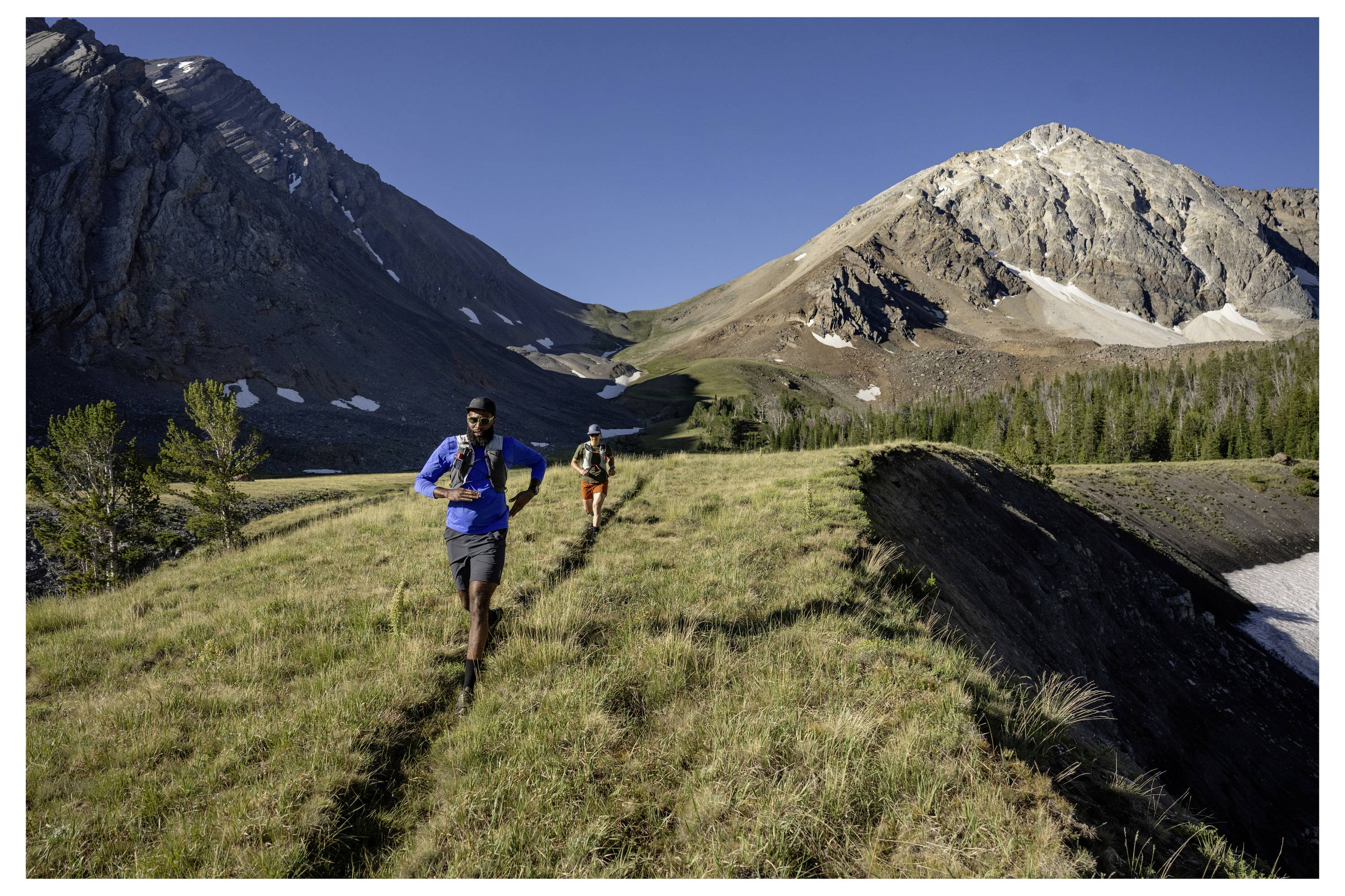 Two people trail running on a grassy mountain path, surrounded by steep, rocky peaks under a clear blue sky.