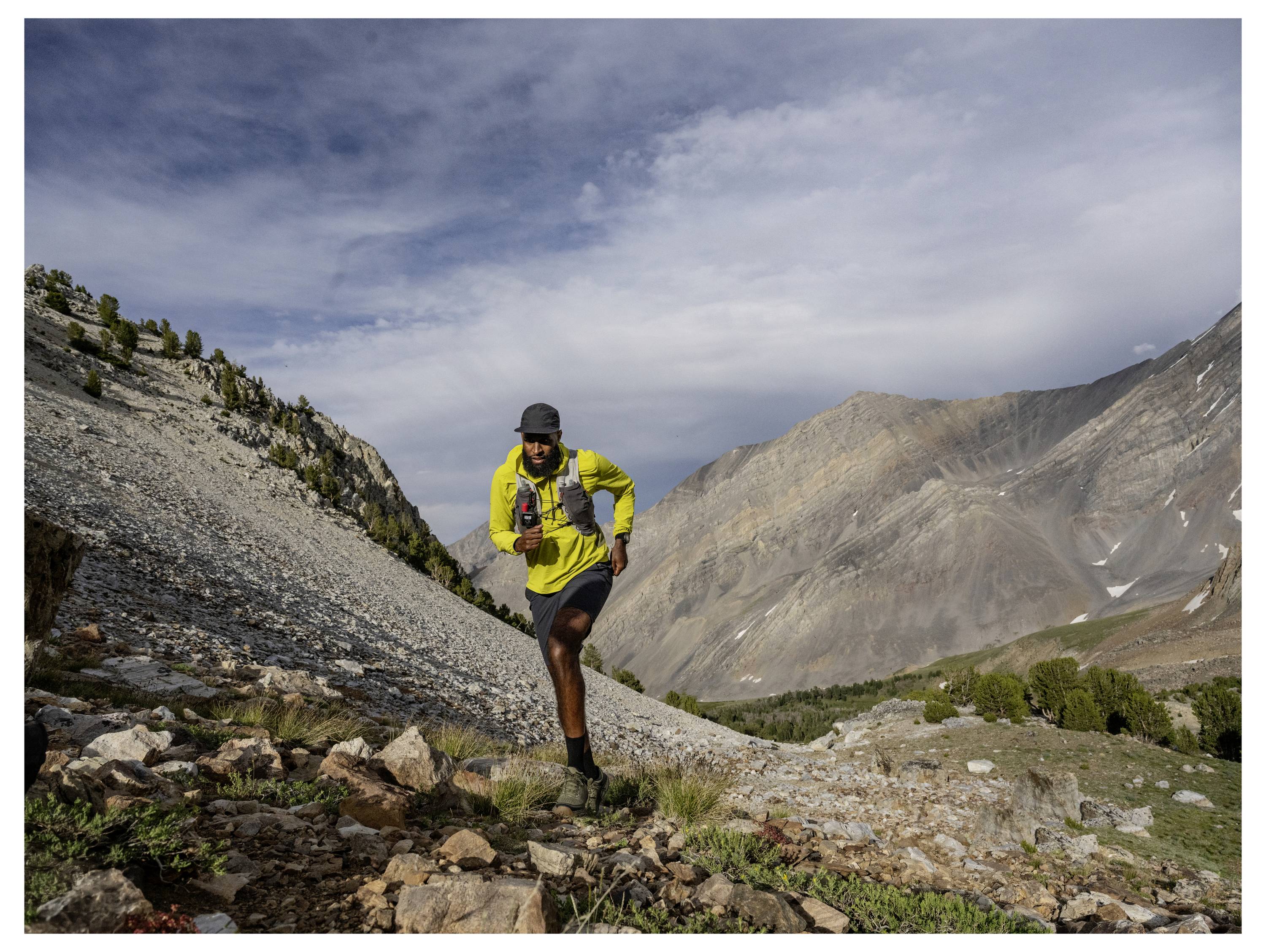 A person running uphill on a rocky mountain trail, wearing a bright yellow shirt and carrying a hydration vest, under a cloudy sky.