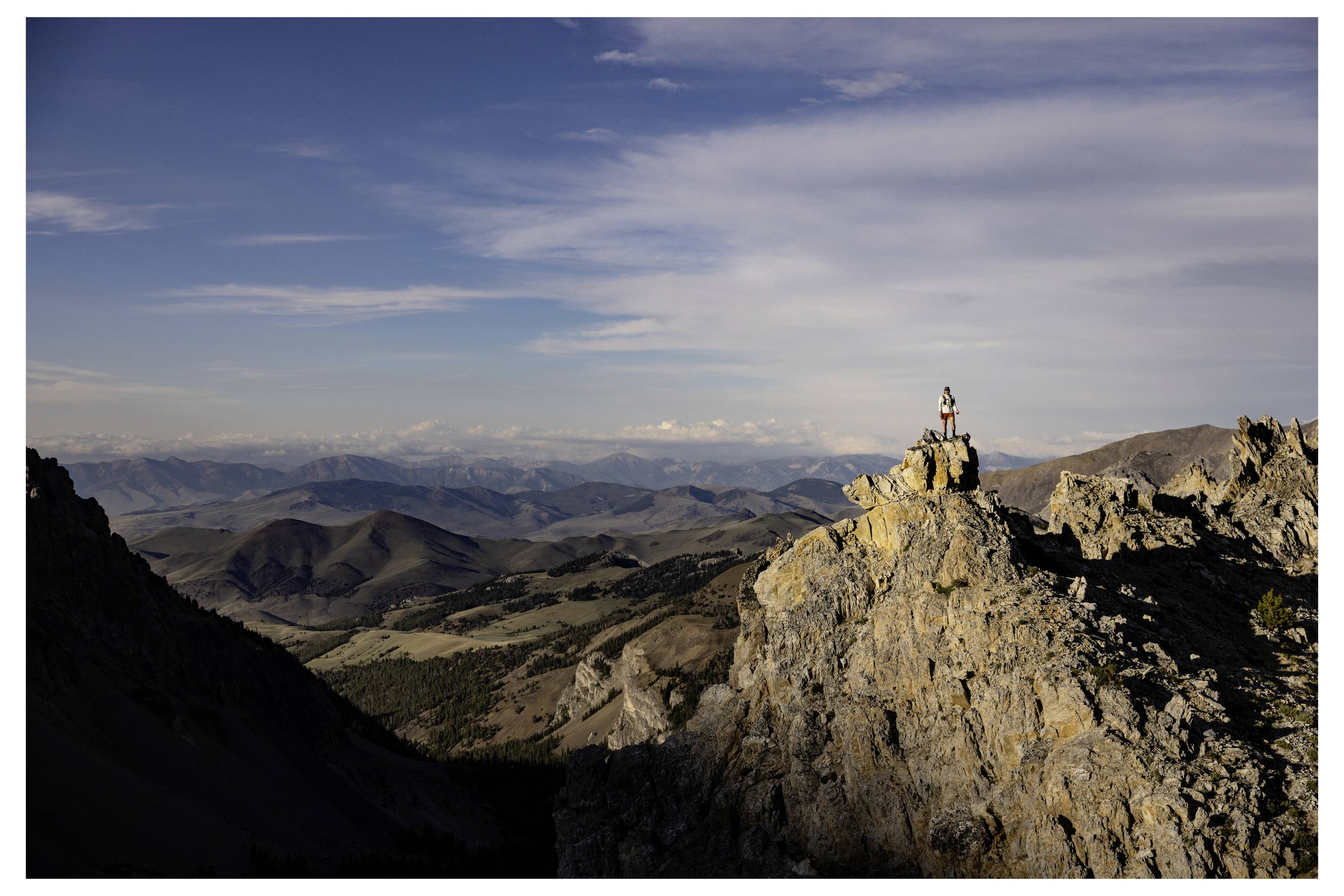 A person stands atop a rocky mountain peak, overlooking a vast landscape of valleys and distant mountains under a blue sky with clouds.
