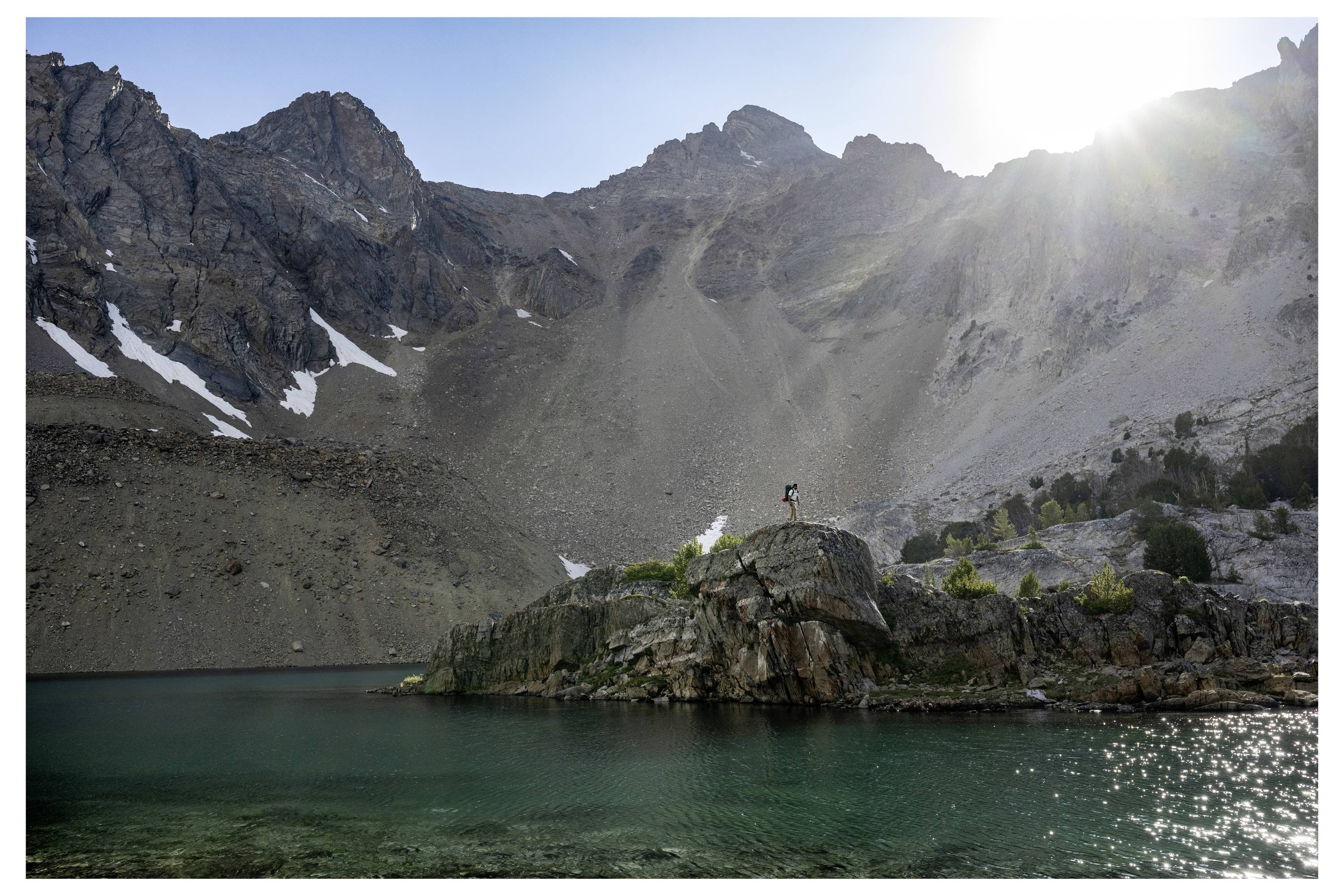 A person stands triumphantly on a rocky outcrop surrounded by a clear green lake and towering mountain peaks under a bright sun.