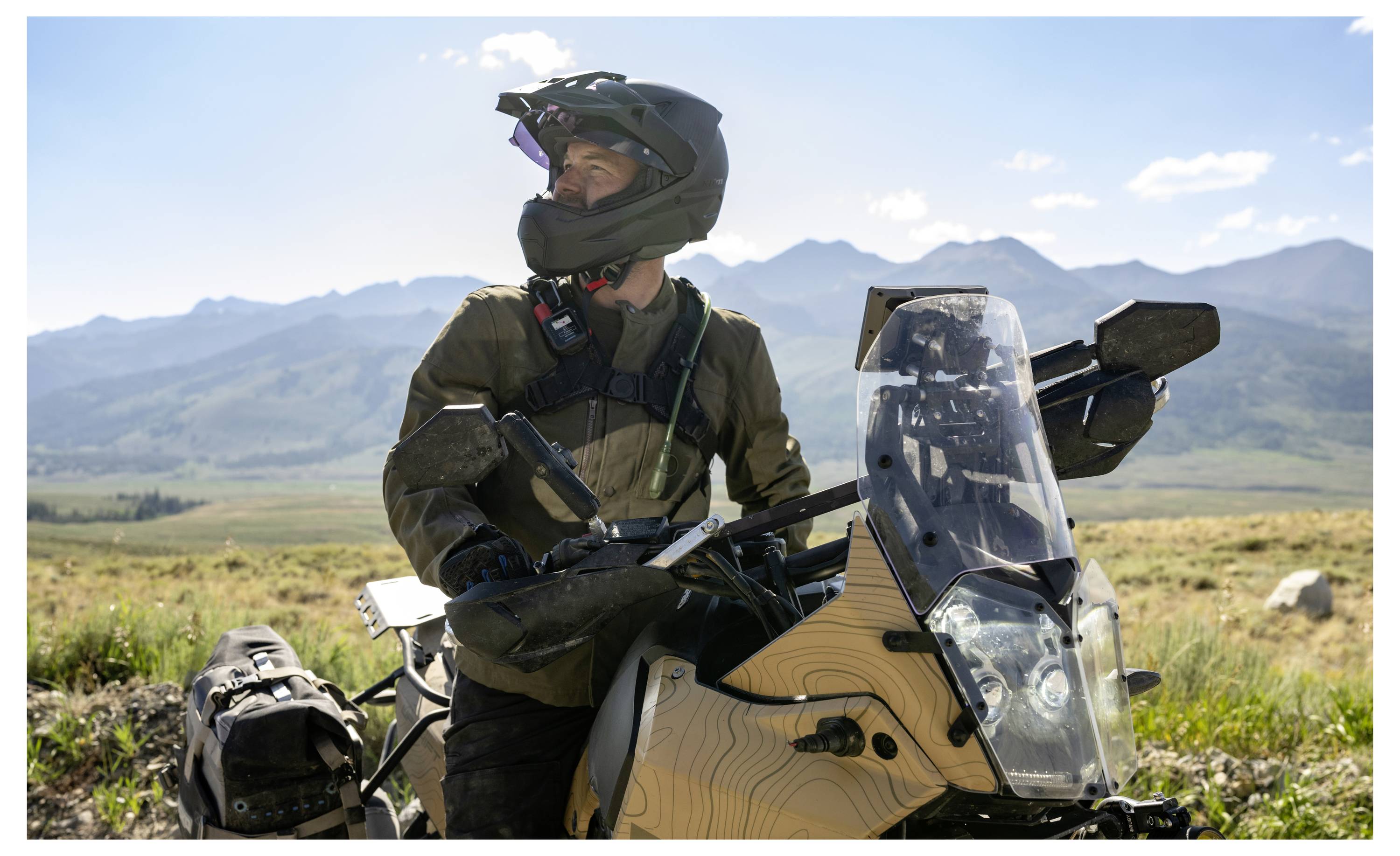 A person in protective gear rides a motorcycle in a mountainous landscape under a clear sky, looking to the side, suggesting exploration.