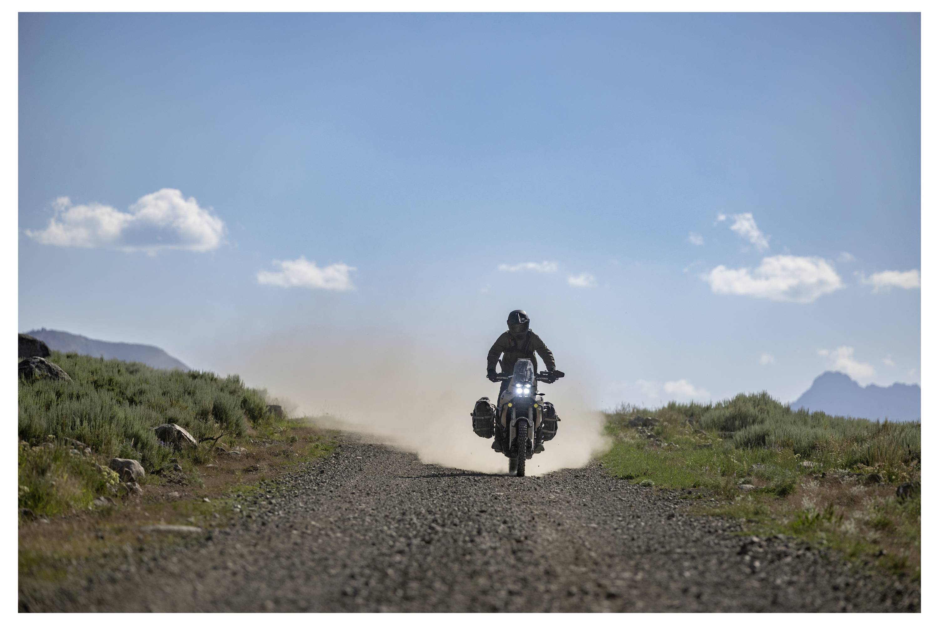 A person rides a motorcycle on a dirt road, surrounded by open landscape with grass and mountains under a clear blue sky with clouds.