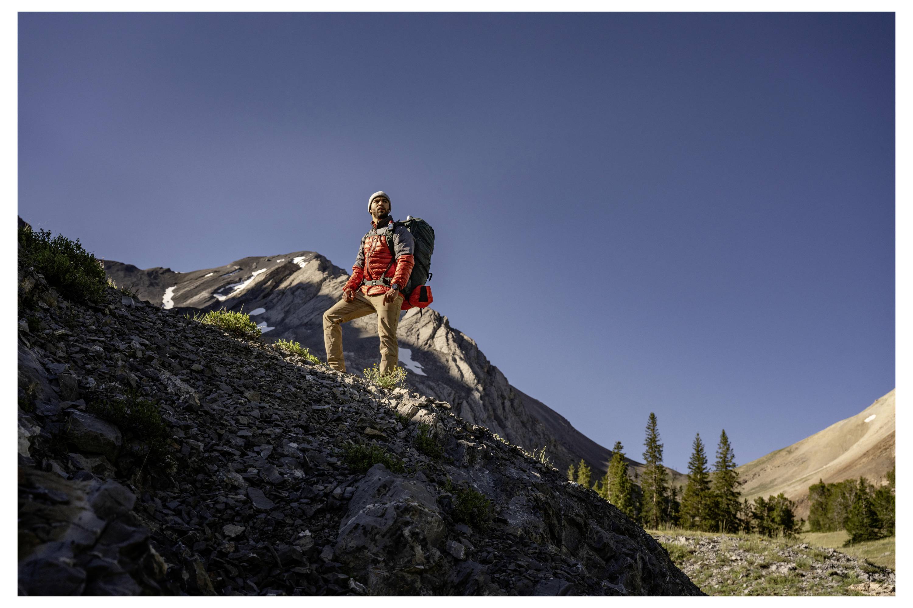 A hiker wearing a backpack stands on a rocky mountain slope under a clear blue sky, with distant trees and peaks in the background.